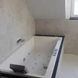 Nowoczesna wanna z hydromasażem w łazience ze ścianami wykończonymi w stylu betonu architektonicznego, widok z boku, w tle okno.