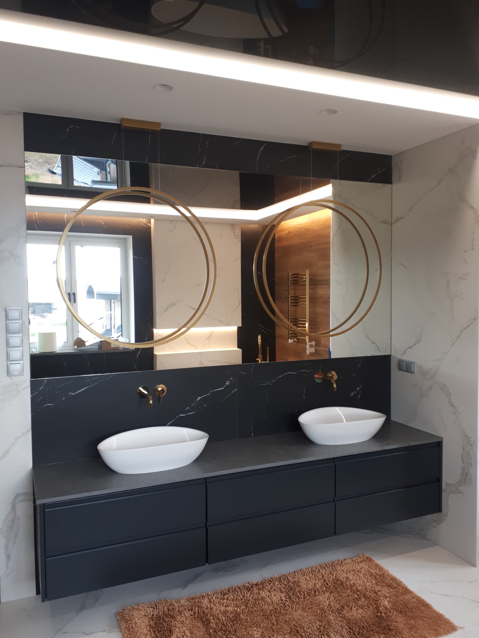 Elegancka łazienka z podwójną umywalką nablatową, czarnym blatem i szafkami, okrągłymi złotymi lampami wiszącymi i lustrem na czarnej ścianie z marmurowym wzorem.