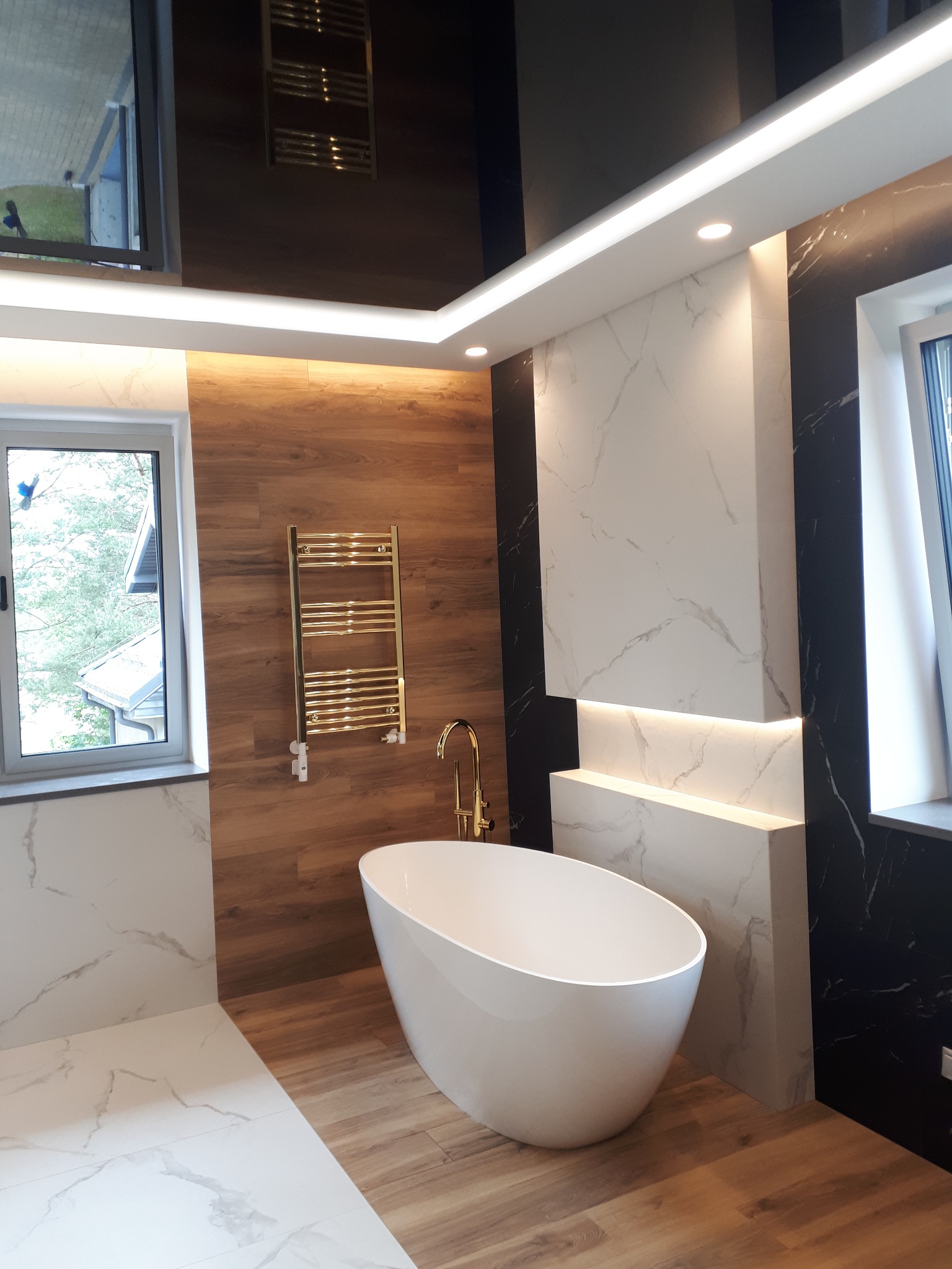 Nowoczesna łazienka z wolnostojącą wanną, złotą armaturą i grzejnikiem drabinkowym na tle drewnopodobnych płytek, marmurowe panele ścienne z podświetleniem LED, okno z widokiem na zewnątrz.