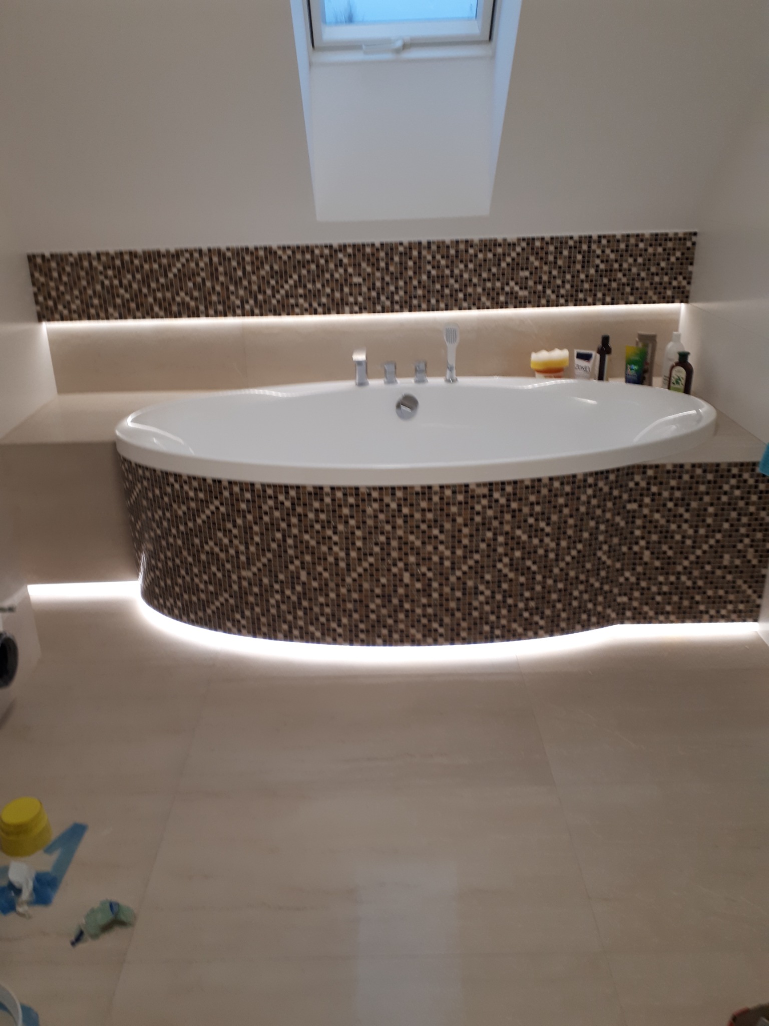 Nowoczesna, owalna wanna obudowana mozaiką w odcieniach brązu, podświetlona taśmą LED, umieszczona w łazience na poddaszu z oknem dachowym. Na wannie widoczne kosmetyki.