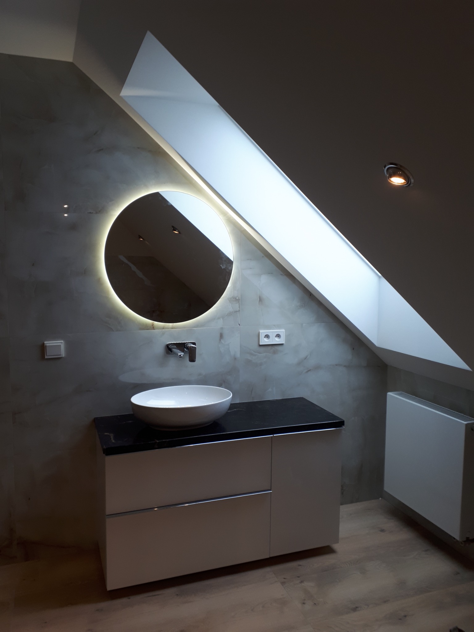 Wykończona łazienka na poddaszu ze skośnym oknem dachowym, okrągłym lustrem z podświetleniem LED, umywalką nablatową na szafce z czarnym blatem i grzejnikiem.