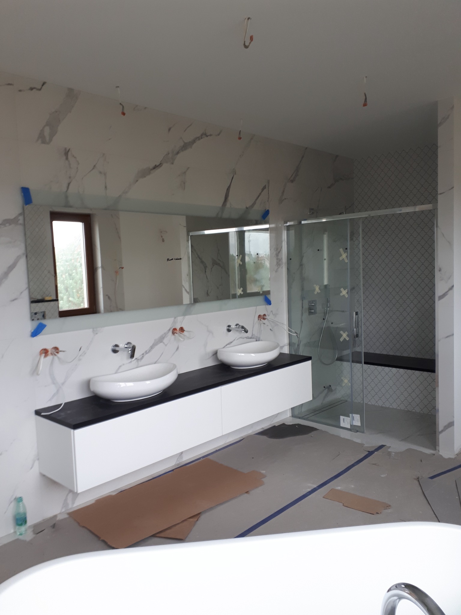 Łazienka w trakcie prac wykończeniowych: umywalki nablatowe, lustro z niebieską taśmą, kabina prysznicowa z mozaiką, widoczne przewody i zabezpieczenia na podłodze.