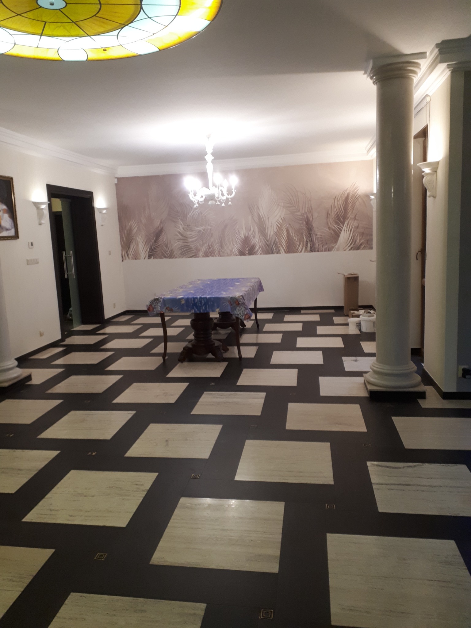 Przestronne wnętrze z dekoracyjną kolumną, wzorzystą tapetą w odcieniach beżu i brązu, oraz podłogą z kwadratowych płytek w kontrastujących kolorach. Na suficie widoczny witraż i żyrandol.