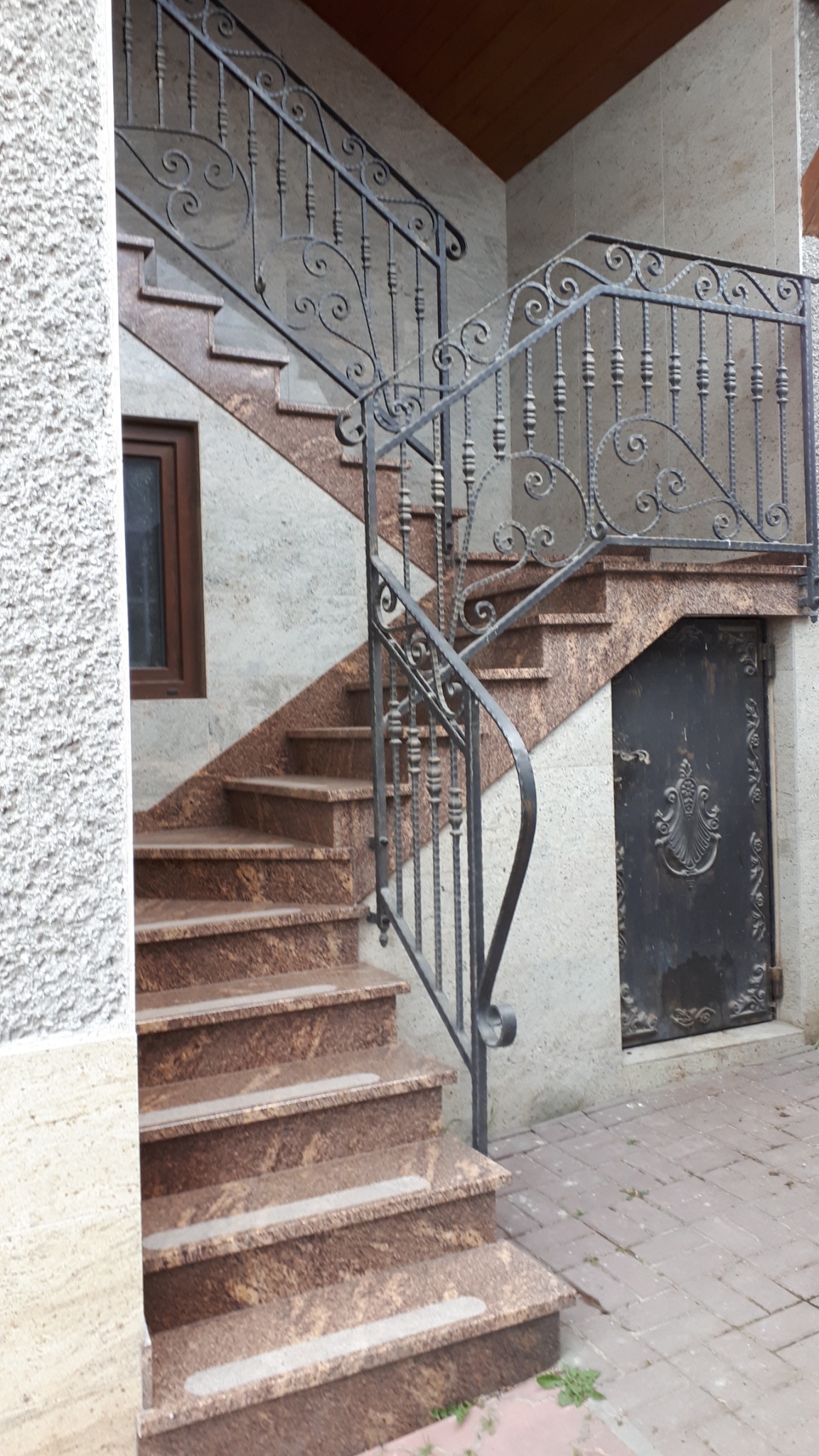 Zewnętrzne schody z brązowego granitu z ozdobną, kutą balustradą, prowadzące do drzwi piwnicznych z dekoracyjnym ornamentem.