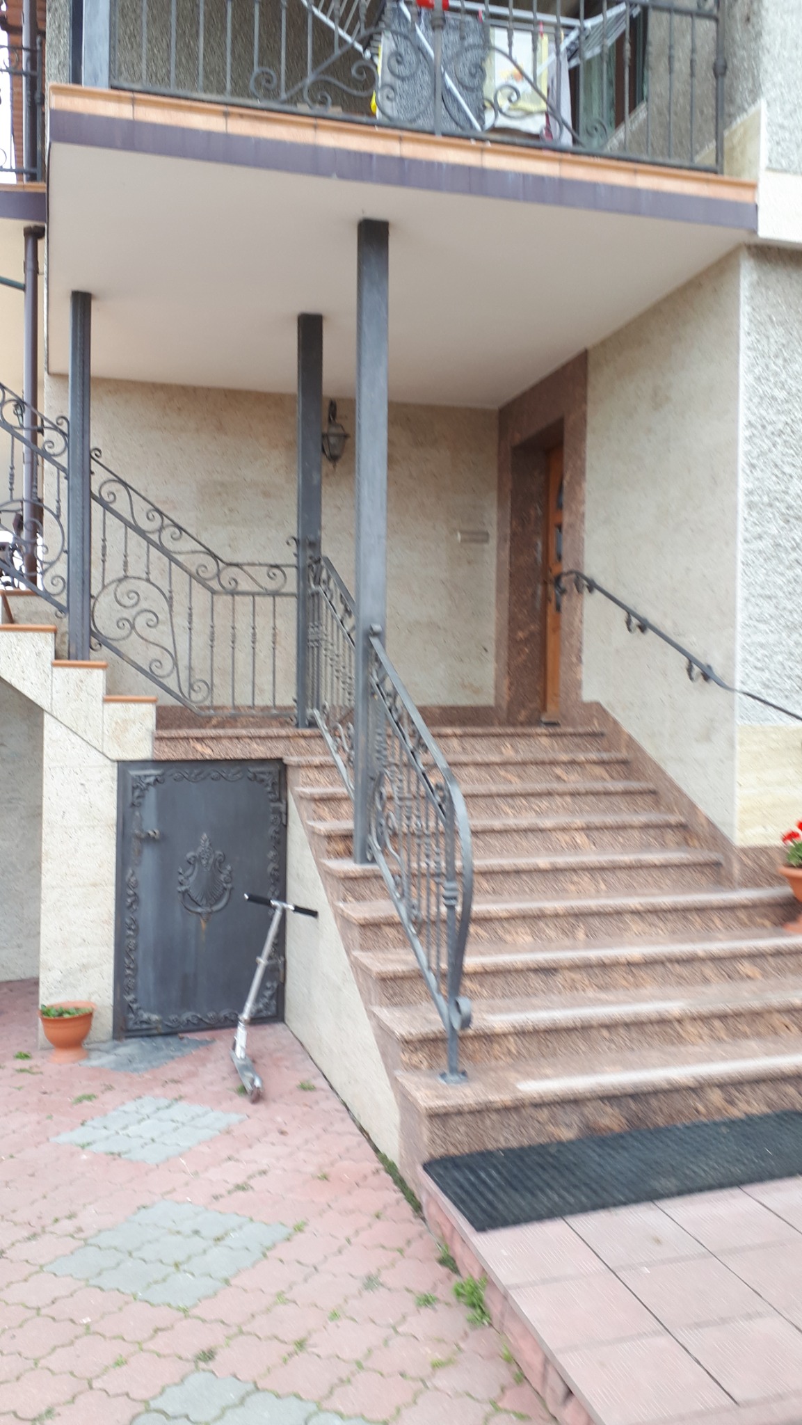 Zewnętrzne schody wejściowe z brązowego granitu prowadzące do drzwi wejściowych, z ozdobną, kutą balustradą i metalowymi słupami, na dole po lewej stronie ciemne drzwi piwniczne z dekoracyjnym...