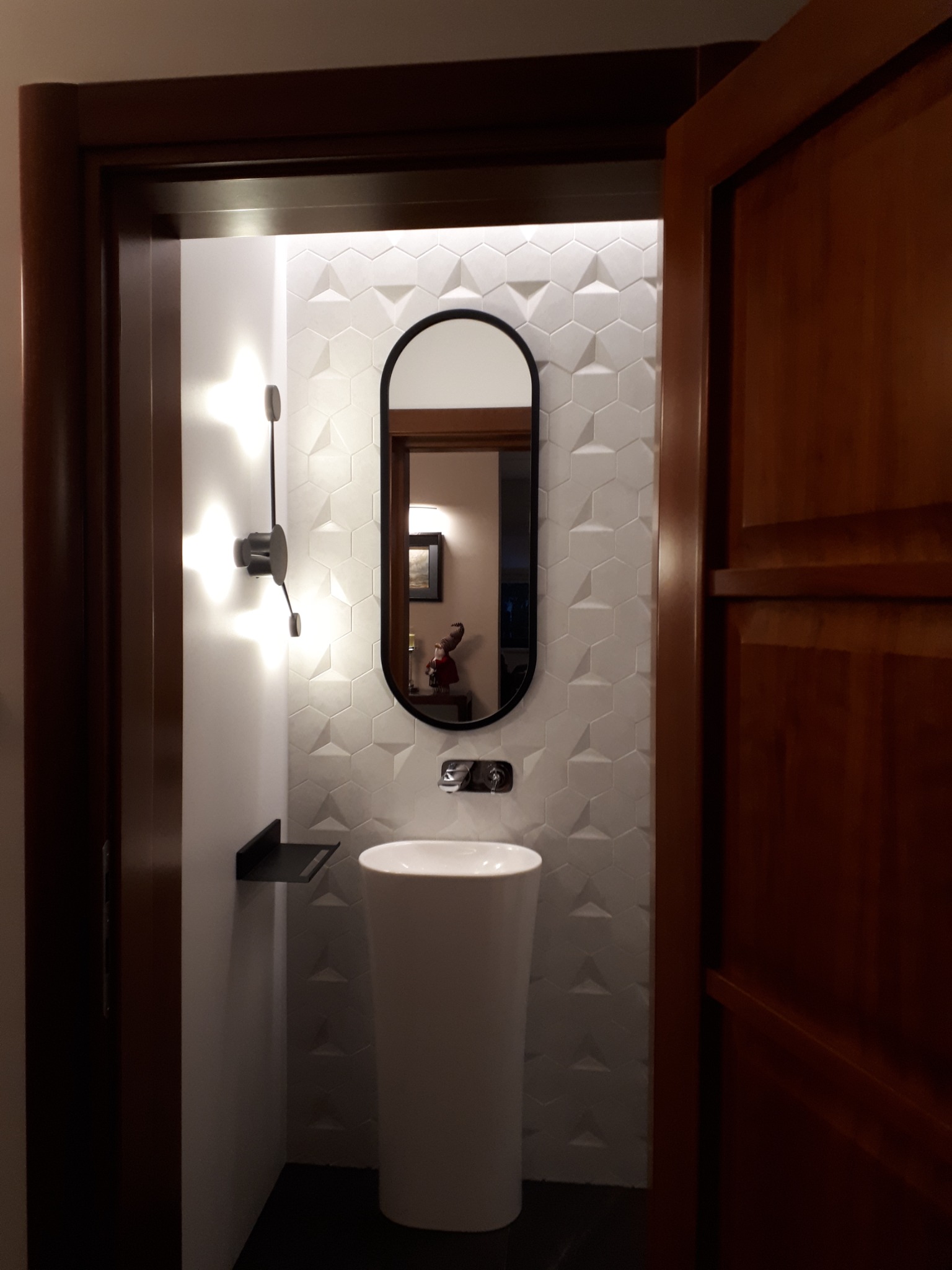 Nowoczesna łazienka z umywalką kolumnową i lustrem w czarnej ramie na tle ściany z białych trójwymiarowych płytek heksagonalnych, oświetlona designerskim kinkietem.