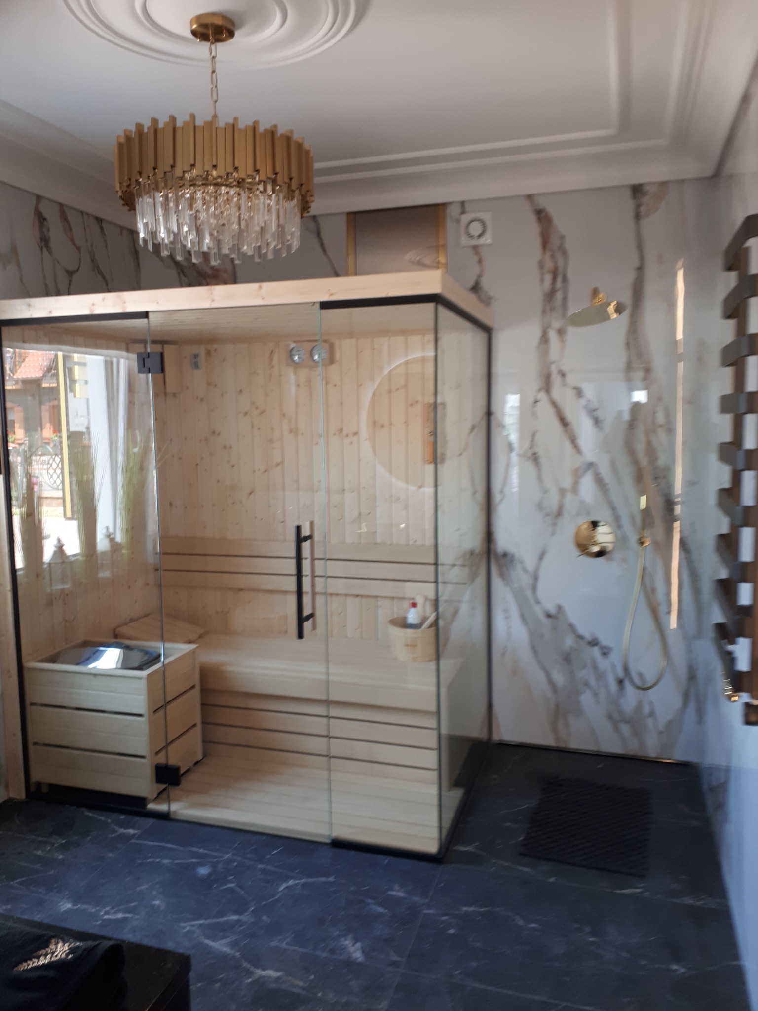 Nowoczesna łazienka z przeszkloną sauną, marmurowe ściany, złota armatura i designerski żyrandol.