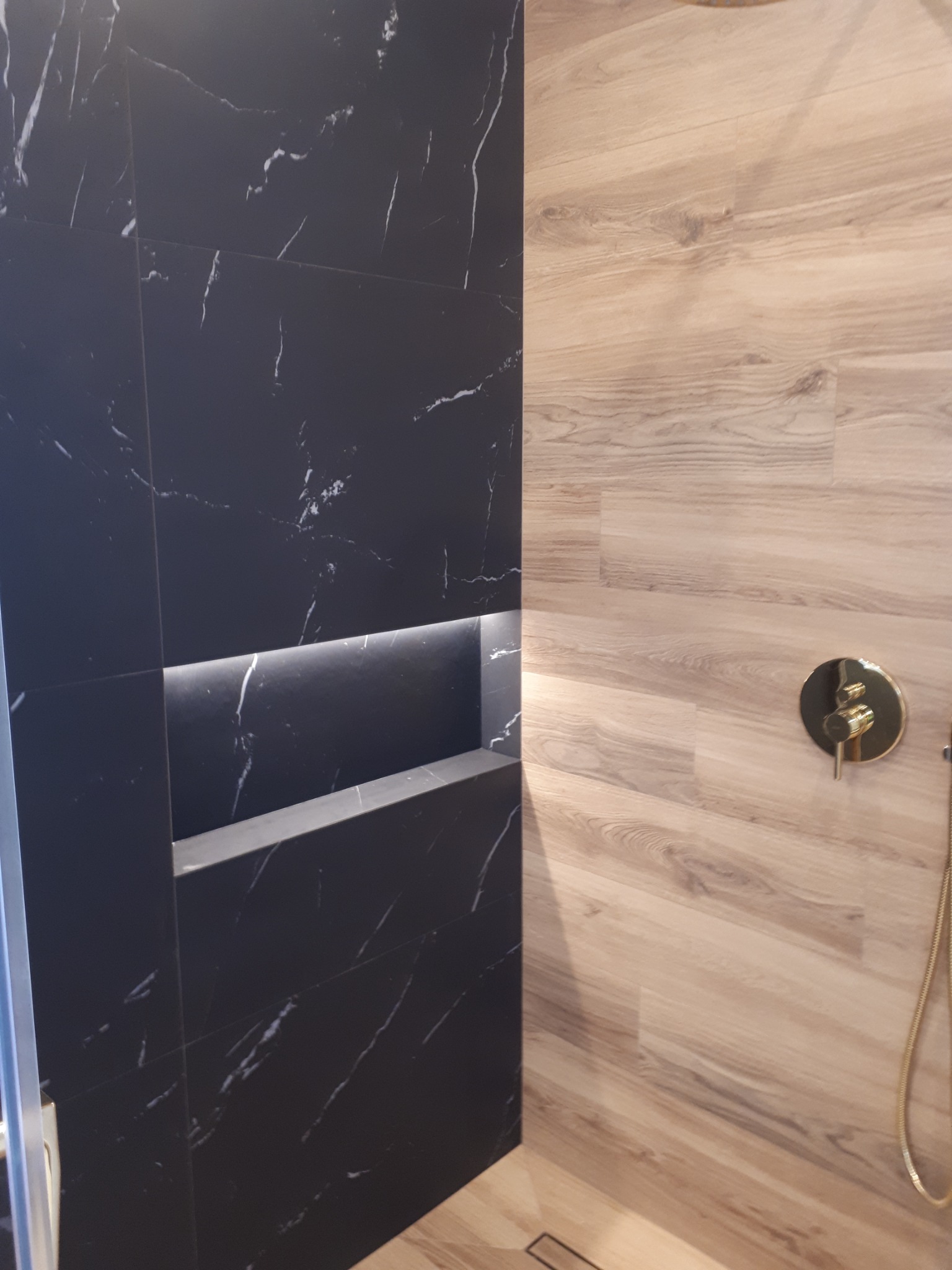 Nowoczesna kabina prysznicowa z czarnymi płytkami marmuropodobnymi z podświetlaną wnęką i drewnopodobnymi panelami ściennymi oraz złotą armaturą.