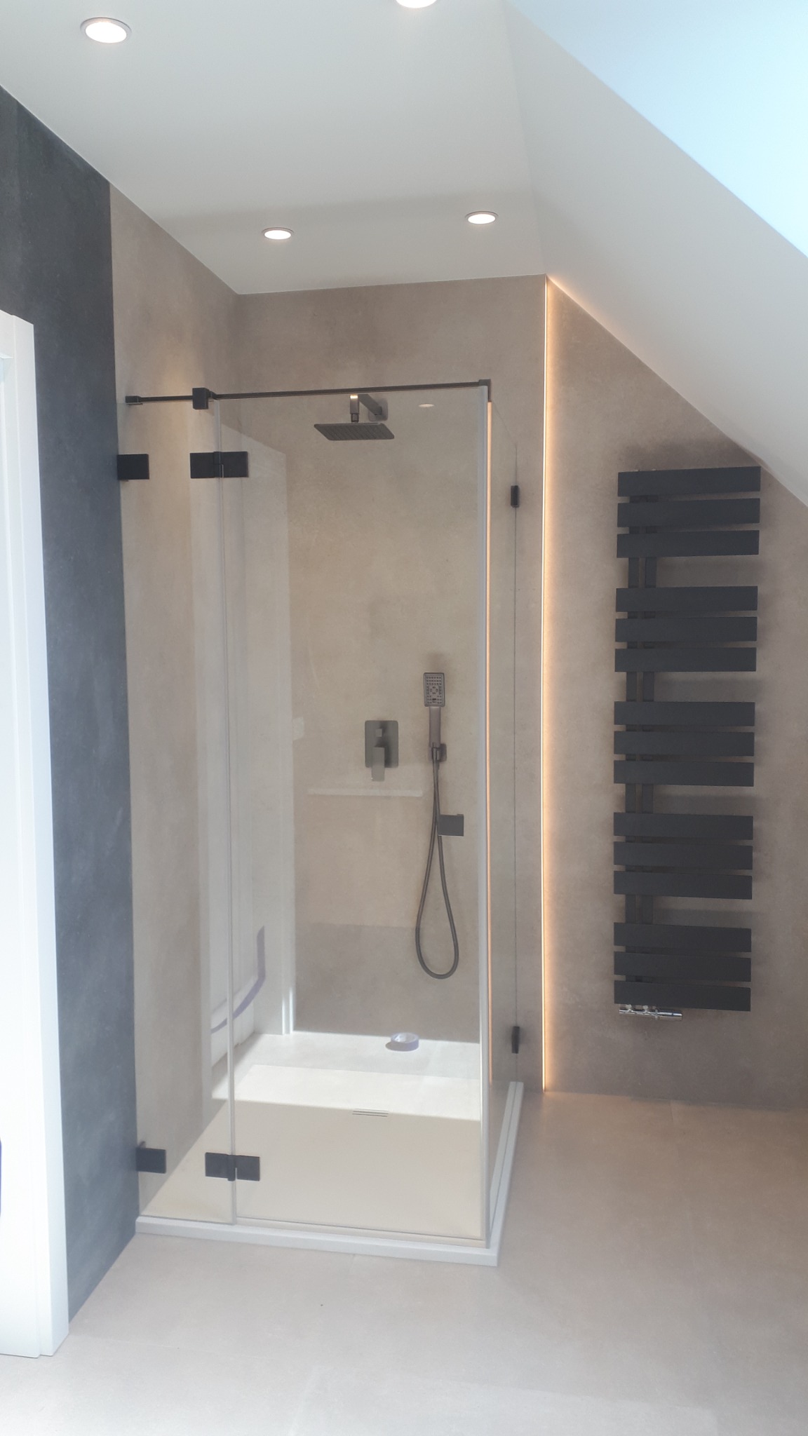 Nowoczesna kabina prysznicowa z czarnymi okuciami i deszczownicą, oświetlona listwą LED, obok grzejnik drabinkowy w łazience ze skośnym sufitem.