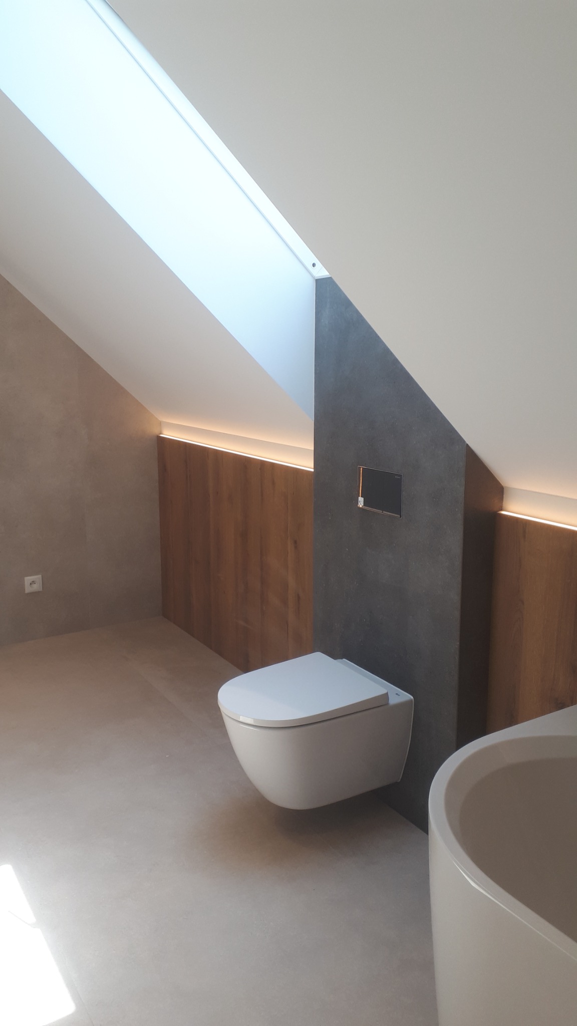 Nowoczesna łazienka na poddaszu ze skośnym sufitem i oknem dachowym, wisząca toaleta na tle szarej ściany, drewniana boazeria z podświetleniem LED, minimalistyczna wanna.