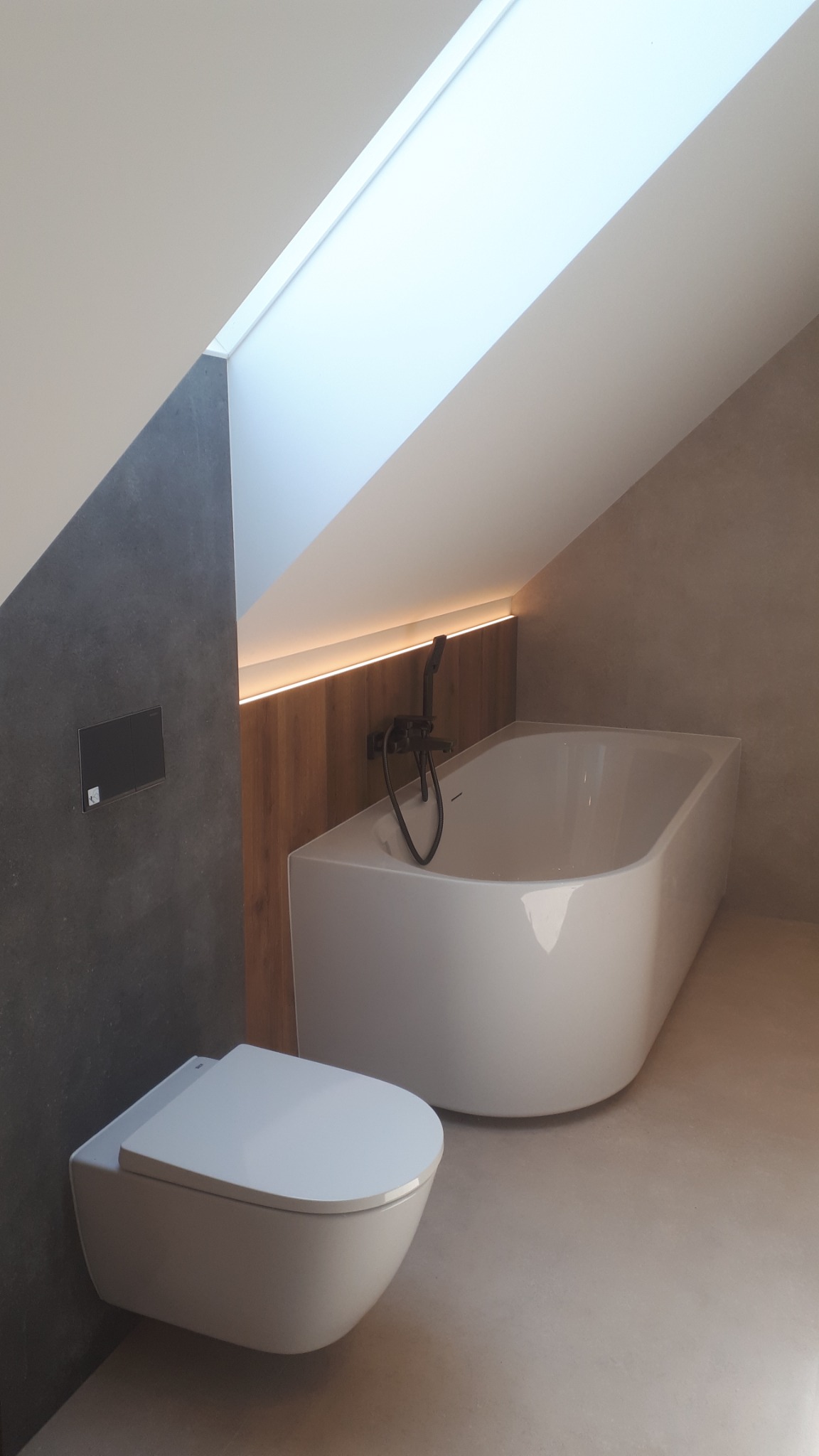 Nowoczesna łazienka na poddaszu ze skośnym sufitem, biała wanna z czarną armaturą, podwieszana toaleta, szara ściana i oświetlenie LED za drewnianą boazerią.