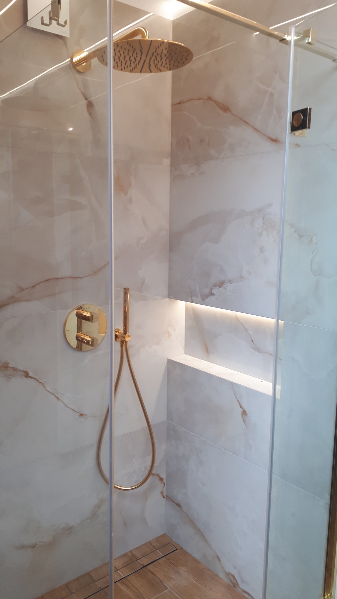 Nowoczesna kabina prysznicowa z deszczownicą i baterią w kolorze złotym, marmurowe płytki na ścianach, podświetlana wnęka, drewnopodobna podłoga z odpływem liniowym.