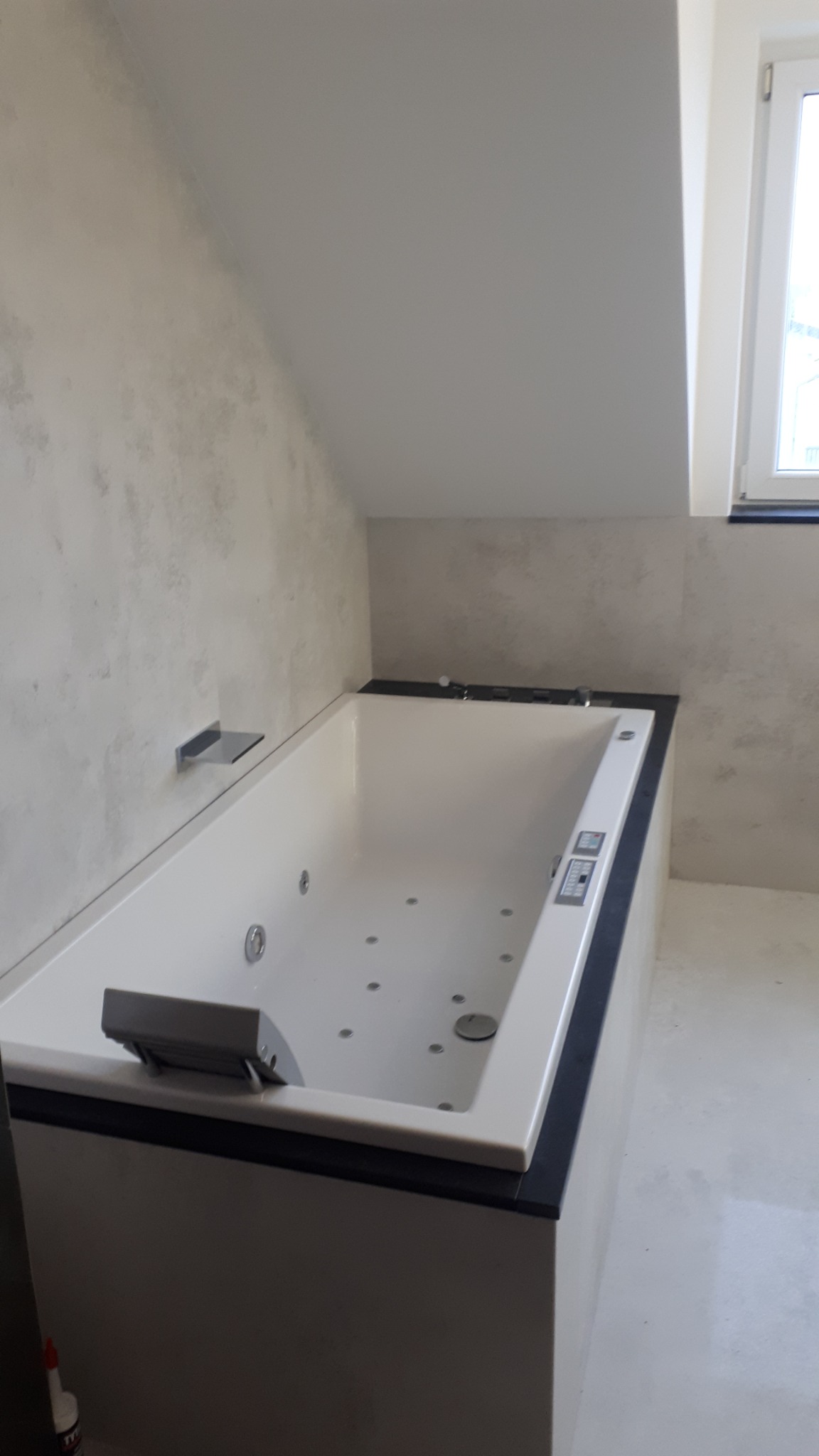 Nowoczesna wanna z hydromasażem w łazience ze ścianami wykończonymi w stylu betonu architektonicznego, widok z boku, w tle okno.