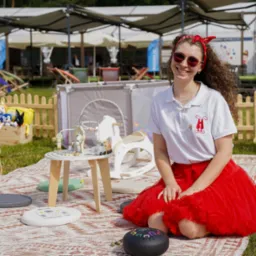 Uśmiechnięta animatorka w czerwonej spódnicy siedzi na kocu piknikowym, otoczona dziecięcymi zabawkami i namiotem tipi. W tle scena z parasolami i leżakami.