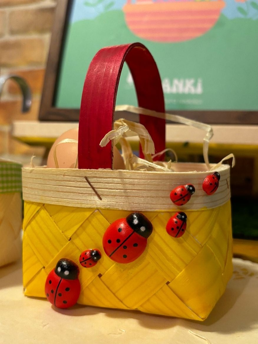 Żółty, pleciony koszyczek wielkanocny z łuby, ozdobiony czerwonymi biedronkami, z czerwonym uchwytem, wypełniony jajkiem i siankiem, na tle plakatu.