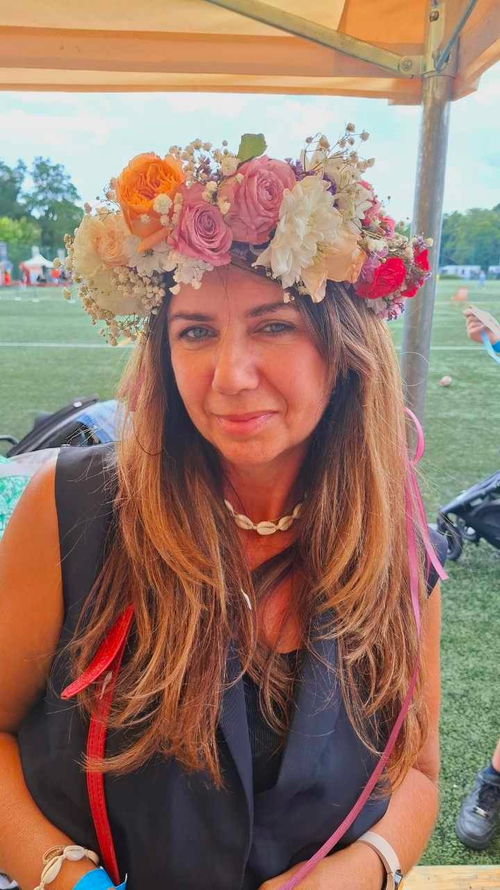 Portret kobiety w wianku z żywych kwiatów (róże, gipsówka, peonie) z różowymi wstążkami na tle zielonej trawy.