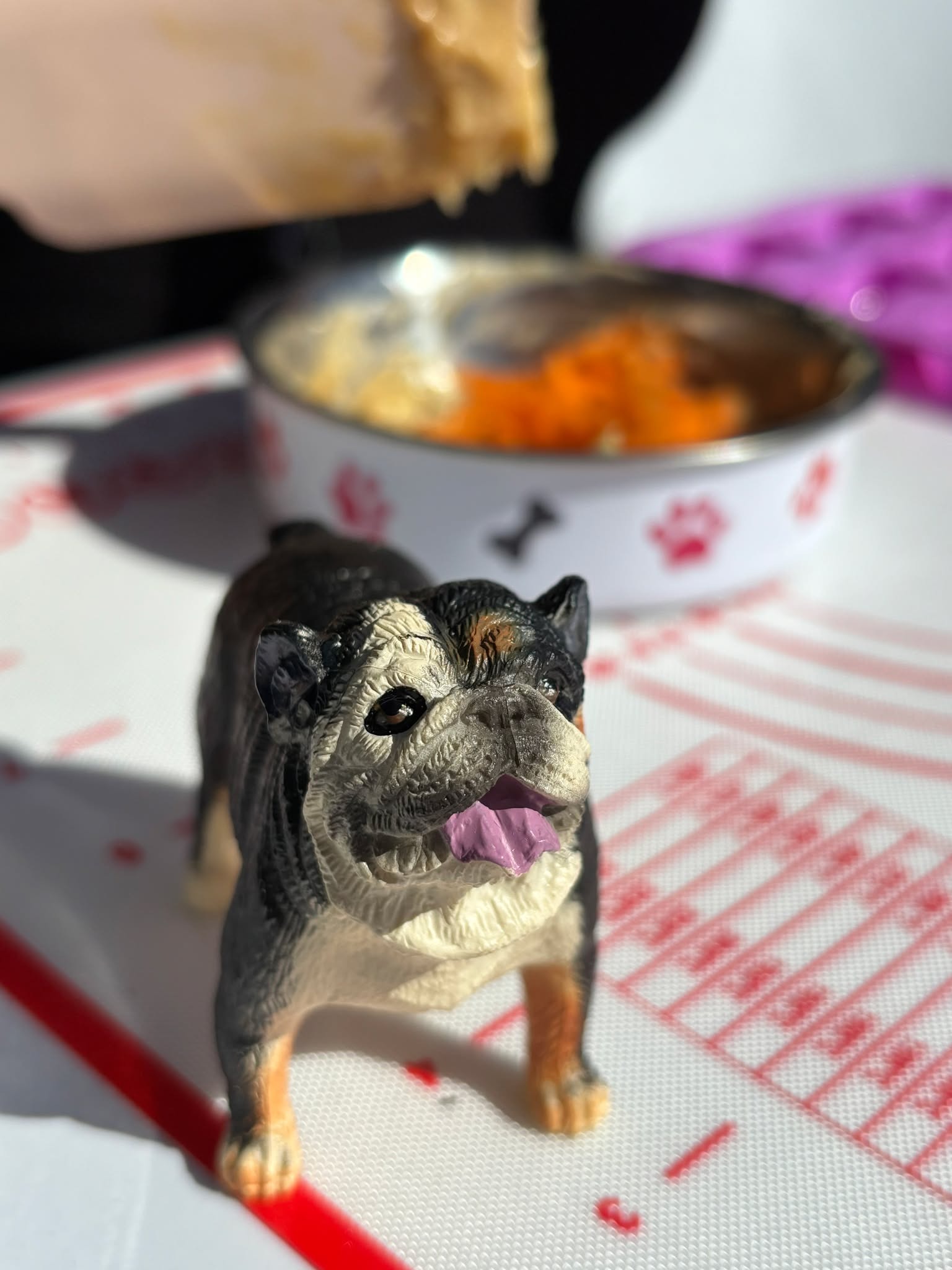 Figurka buldoga na silikonowej macie do pieczenia z miską na karmę w tle, podczas przygotowywania 'psich chrupków', co sugeruje warsztaty kulinarne dla zwierząt.