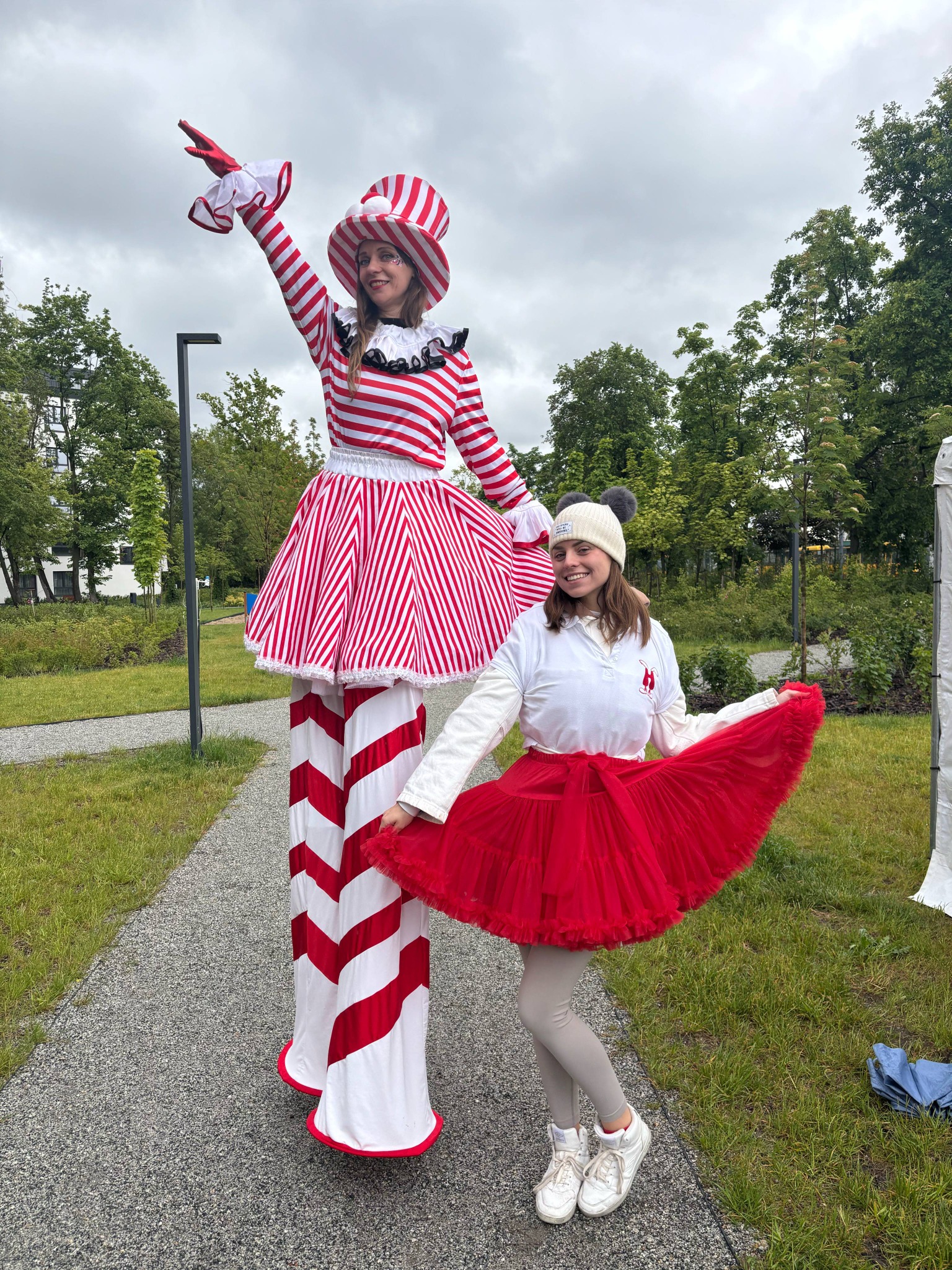 Dwie kobiety w kostiumach: jedna na szczudłach w czerwono-białym stroju, druga w czerwonej spódnicy. W tle park i budynki.