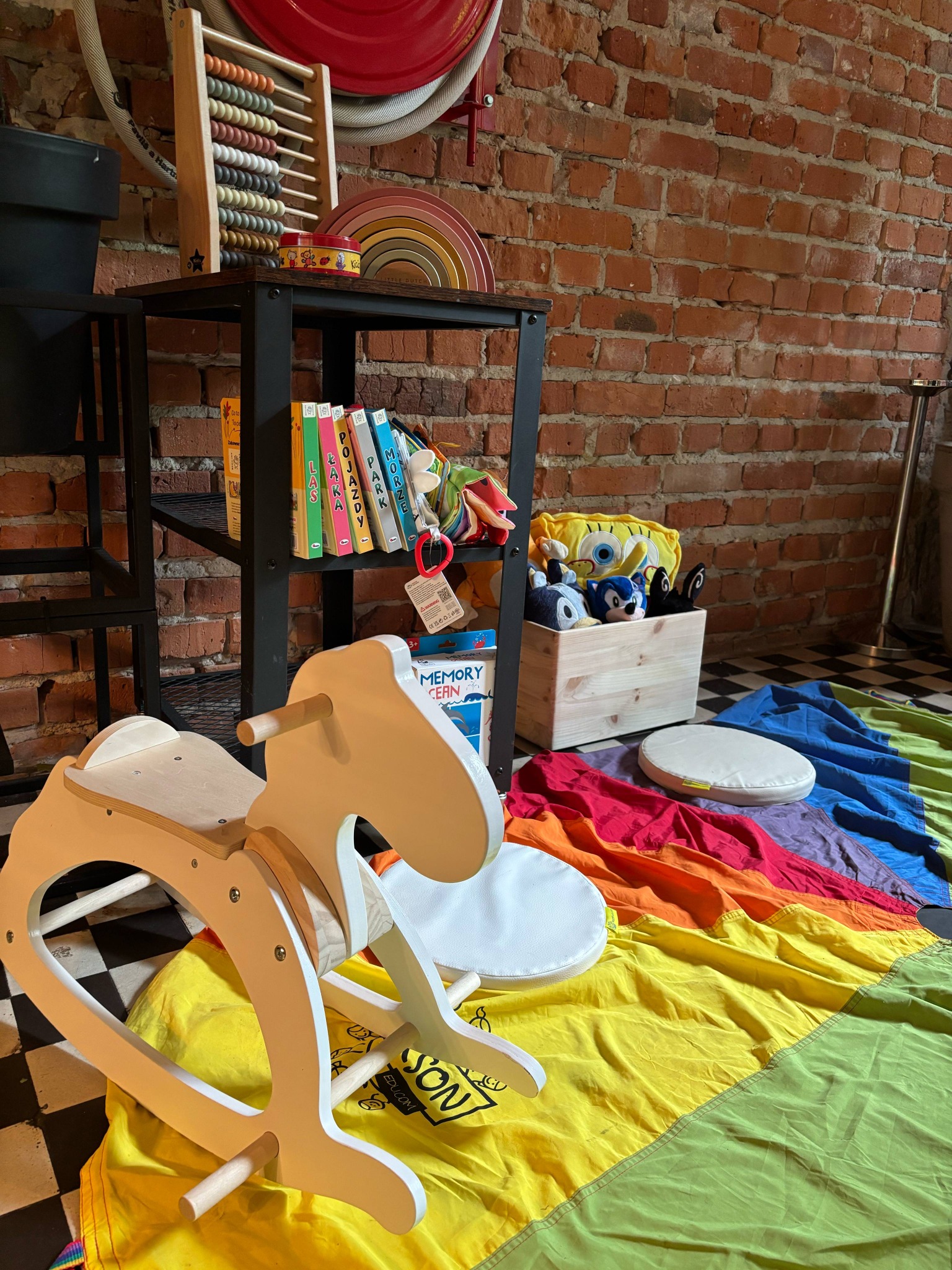 Pokój zabaw dla dzieci z drewnianym koniem na biegunach, kolorową chustą i półką z książkami oraz zabawkami na tle ceglanej ściany.