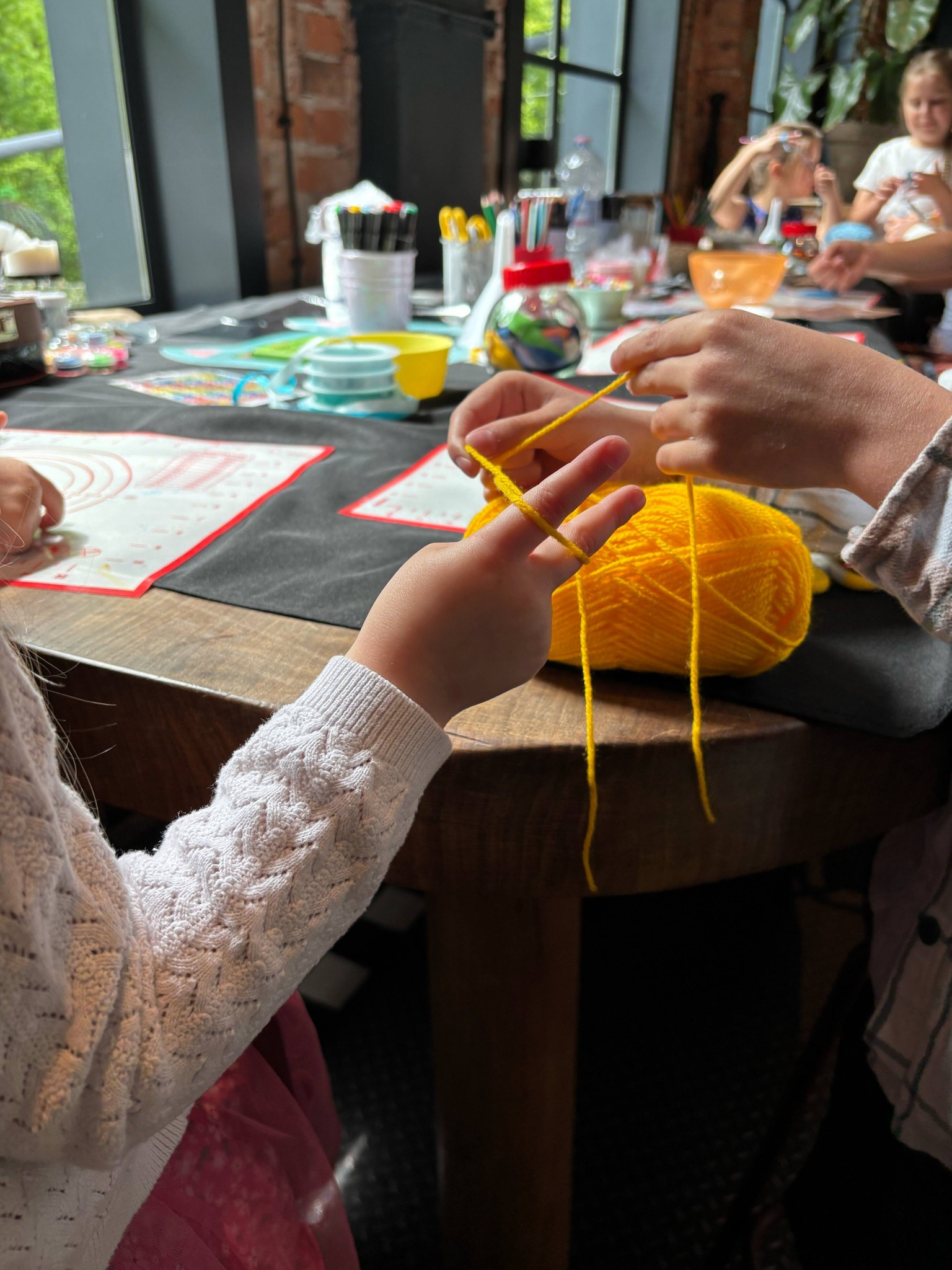Dziecko nawija żółtą włóczkę na palce podczas warsztatów rękodzielniczych przy stole z akcesoriami, w tle widoczne inne dzieci.