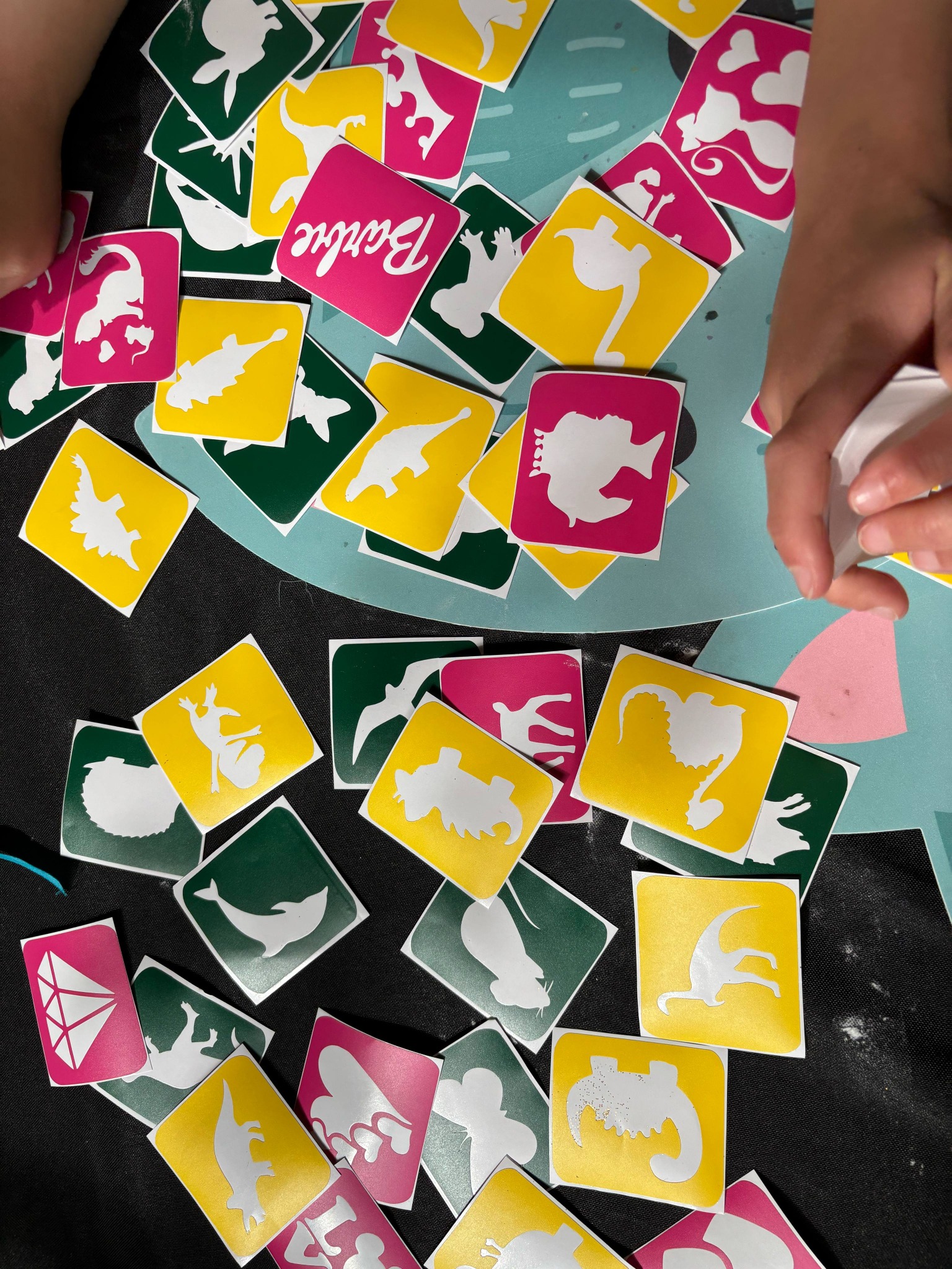 Kolorowe naklejki z różnymi kształtami zwierząt i postaci, rozłożone na stole przez dłonie dziecka. Żółte, zielone i różowe kwadraty z białymi sylwetkami.