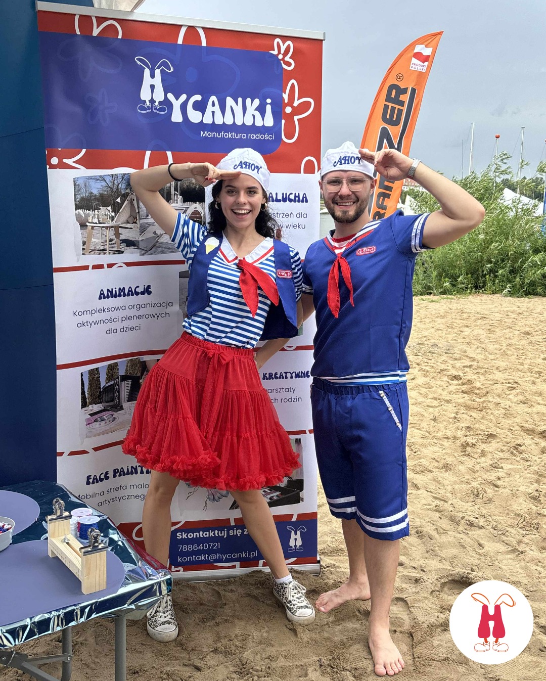 Dwoje animatorów w marynarskich strojach pozuje na plaży przed banerem firmy Hycanki, oferującej animacje dla dzieci. Uśmiechnięci, w czapkach z napisem AHOY.