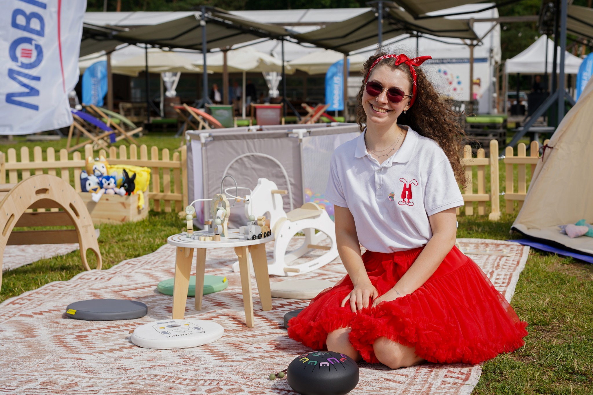 Uśmiechnięta animatorka w czerwonej spódnicy siedzi na kocu piknikowym, otoczona dziecięcymi zabawkami i namiotem tipi. W tle scena z parasolami i leżakami.