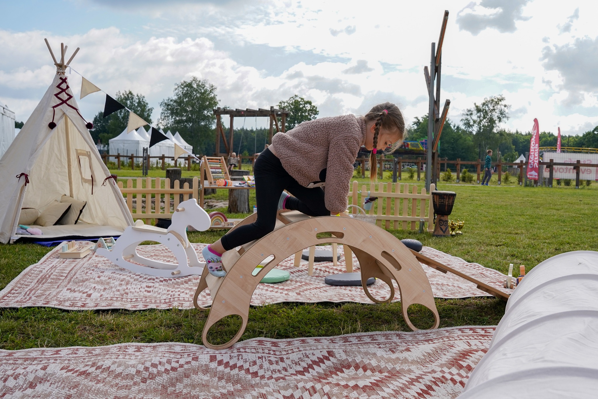 Dziewczynka wspina się na drewniany łuk na pikniku z tipi i zabawkami na trawie. Widoczne huśtawki i płotki, tworzące przestrzeń zabaw na świeżym powietrzu.