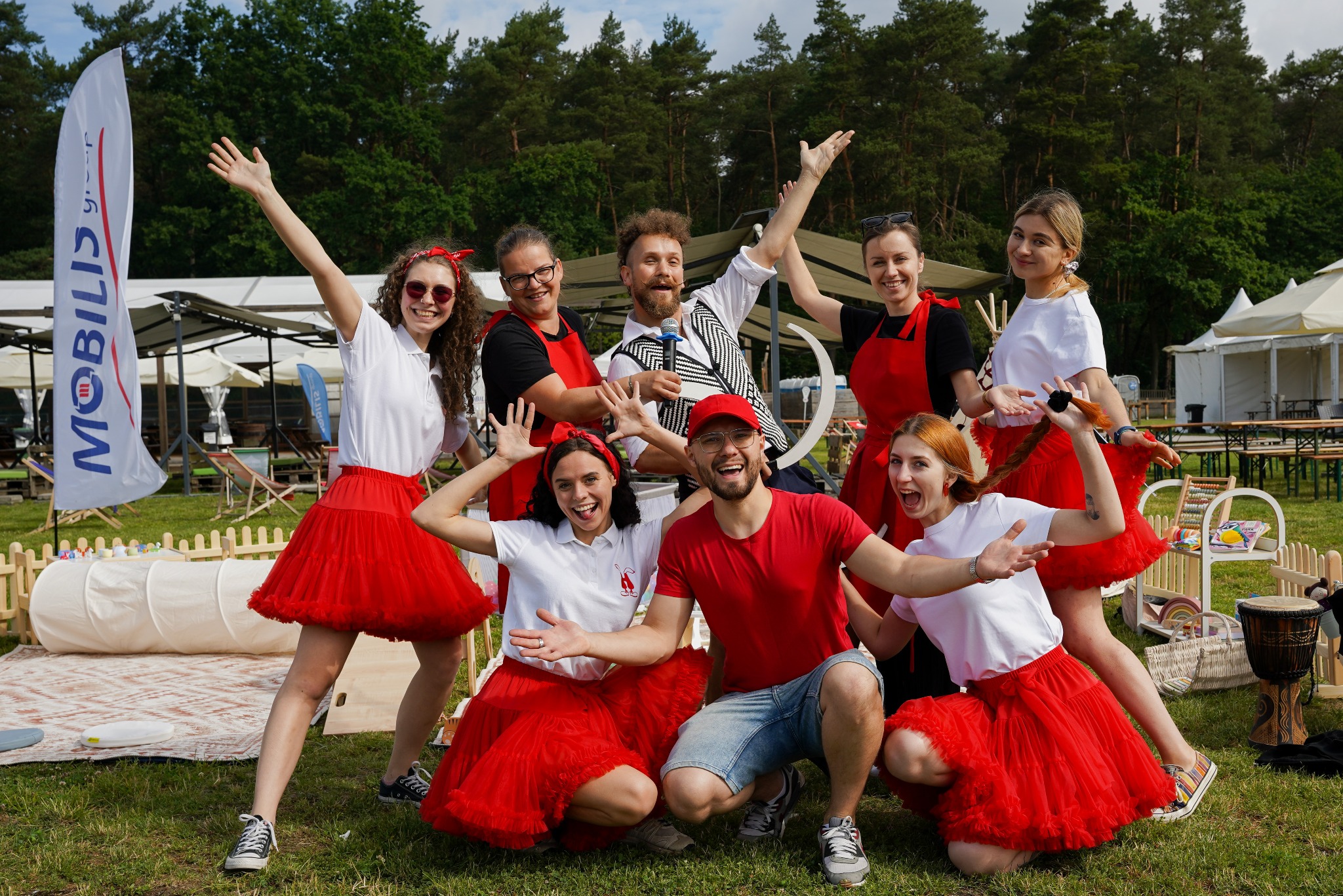Ekipa animatorów w czerwonych tiulowych spódnicach i białych koszulkach pozuje na tle zieleni, z dmuchanym tunelem i flagą reklamową w tle.