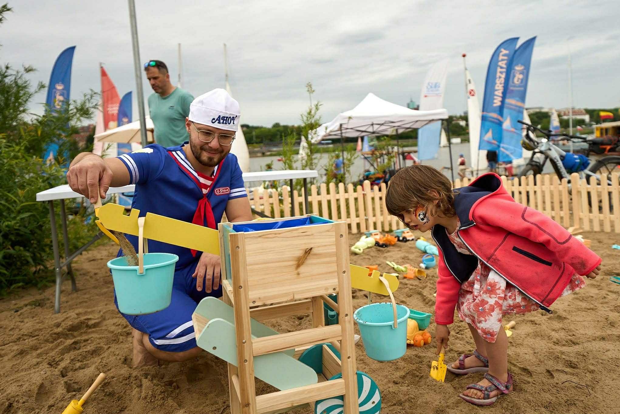Animator w kostiumie marynarza i dziewczynka bawią się w piasku z drewnianą konstrukcją do przesypywania piasku. W tle widoczne flagi i inne osoby.