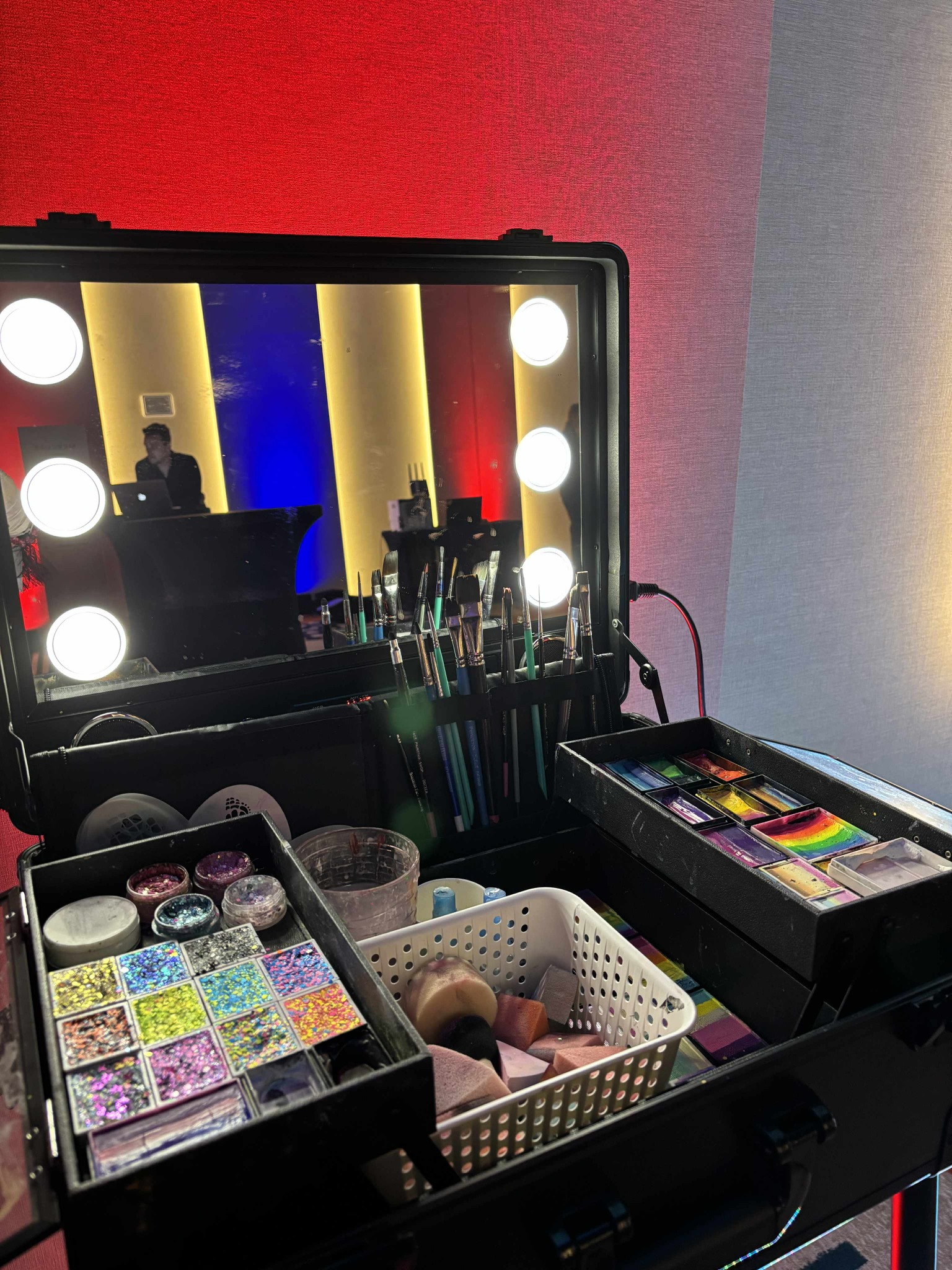 Profesjonalny kufer wizażysty z pędzlami, farbami do twarzy i brokatami. W tle odbicie osoby przy laptopie na scenie z oświetleniem w kolorach flagi.