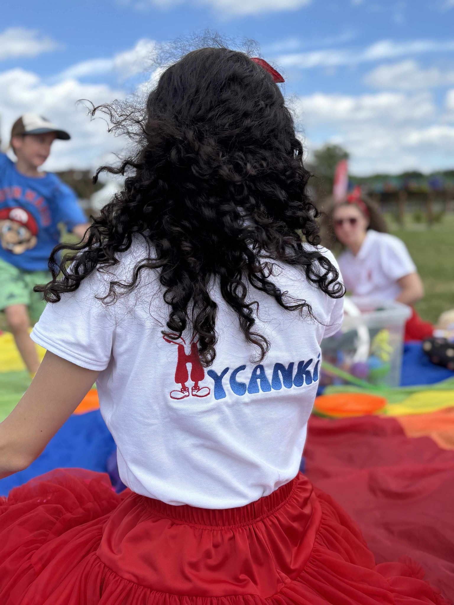 Animator w białej koszulce z logo firmy, w czerwonej spódniczce tiulowej, siedzi na kolorowej chuście animacyjnej podczas eventu plenerowego dla dzieci.