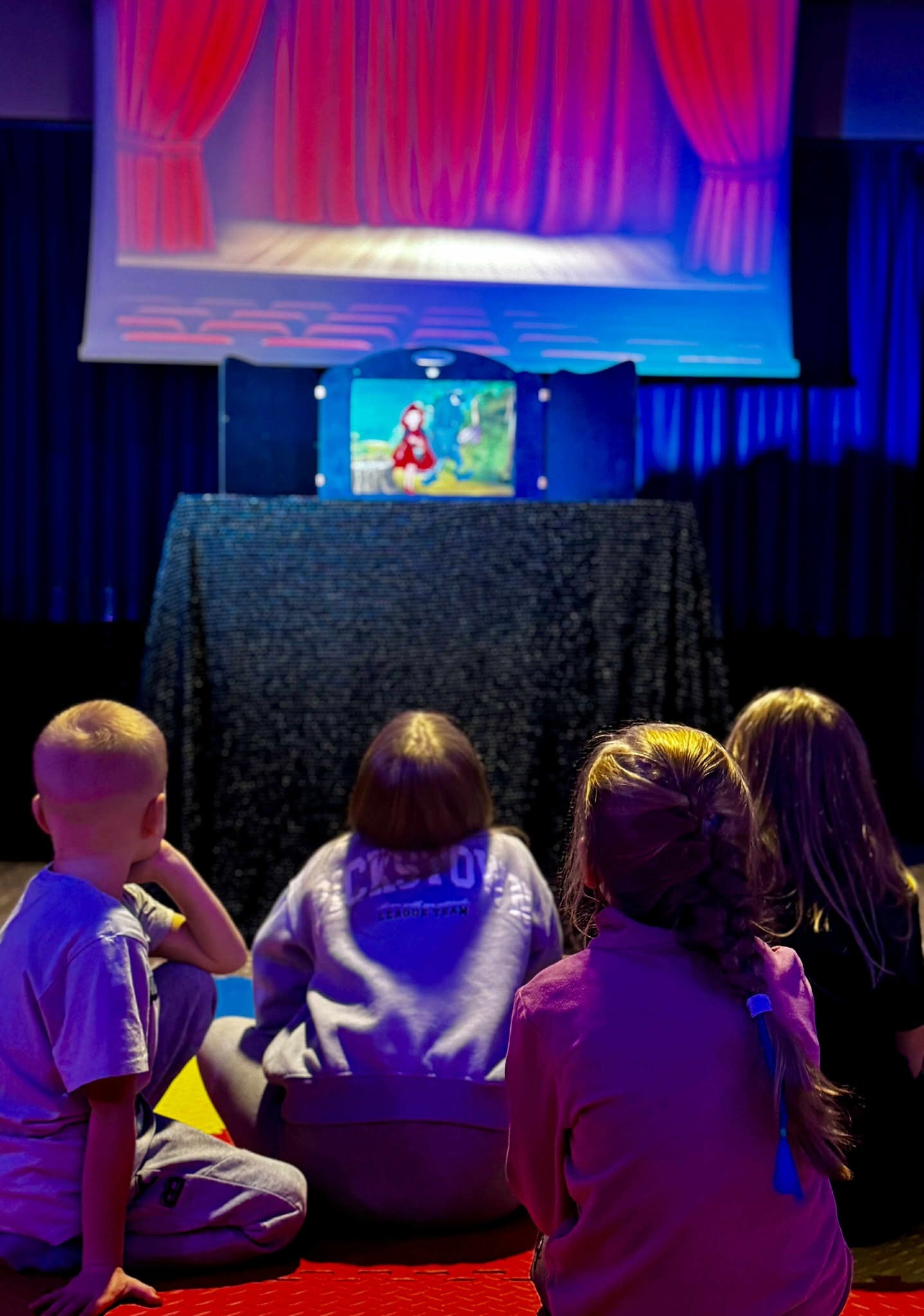 Dzieci siedzą na podłodze, oglądając przedstawienie kukiełkowe z Czerwonym Kapturkiem na małej scenie z czerwonymi zasłonami i niebieskim oświetleniem.