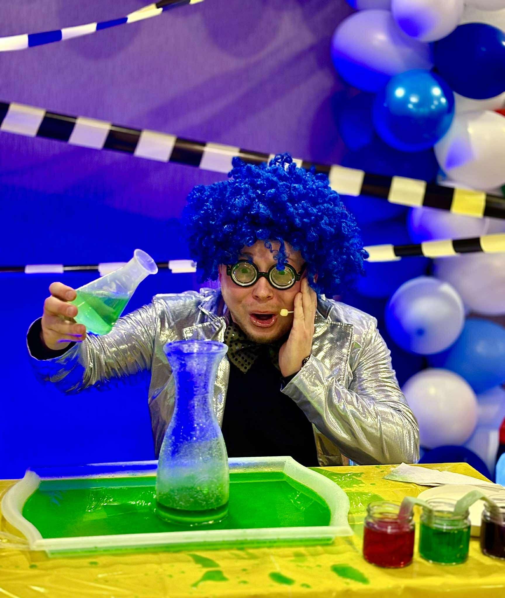 Animator w srebrnej kurtce i niebieskiej peruce w Warszawie prezentuje eksperyment chemiczny z kolorowymi płynami podczas imprezy dla dzieci.