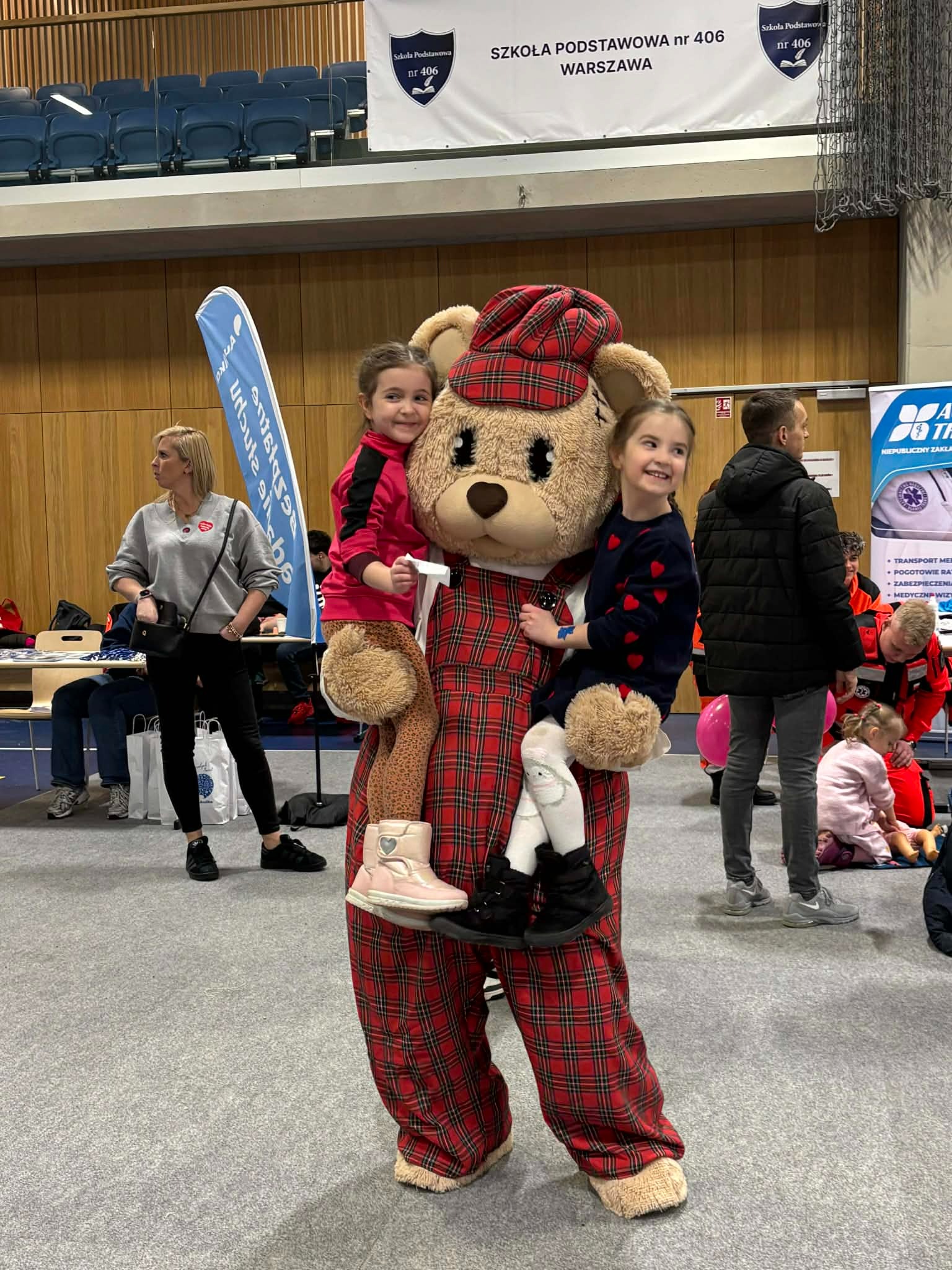 Uśmiechnięty animator w kostiumie pluszowego misia w czerwoną kratę, niosący na rękach dwie dziewczynki podczas wydarzenia w hali sportowej.