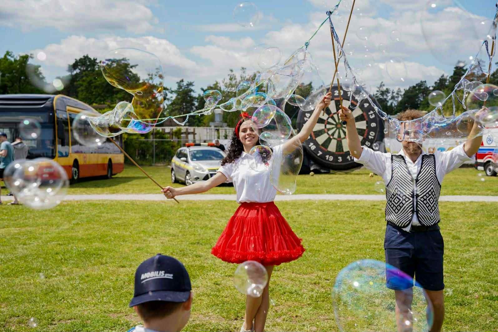 Uśmiechnięta animatorka w czerwonej spódnicy i mężczyzna w kamizelce tworzą gigantyczne bańki mydlane na tle zielonej trawy, autobusu i tarczy do darta.