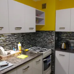 Nowoczesna kuchnia z białymi szafkami, mozaikową ścianą nad blatem i intensywnie żółtą ścianą w tle, widok na zlewozmywak, kuchenkę i akcesoria kuchenne.