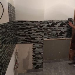 Regameh - Montaż mozaiki z drobnych, poziomych płytek w odcieniach szarości i zieleni na ścianie łazienki, widoczne fragmenty instalacji hydraulicznej i niedokończone fragmenty ściany.