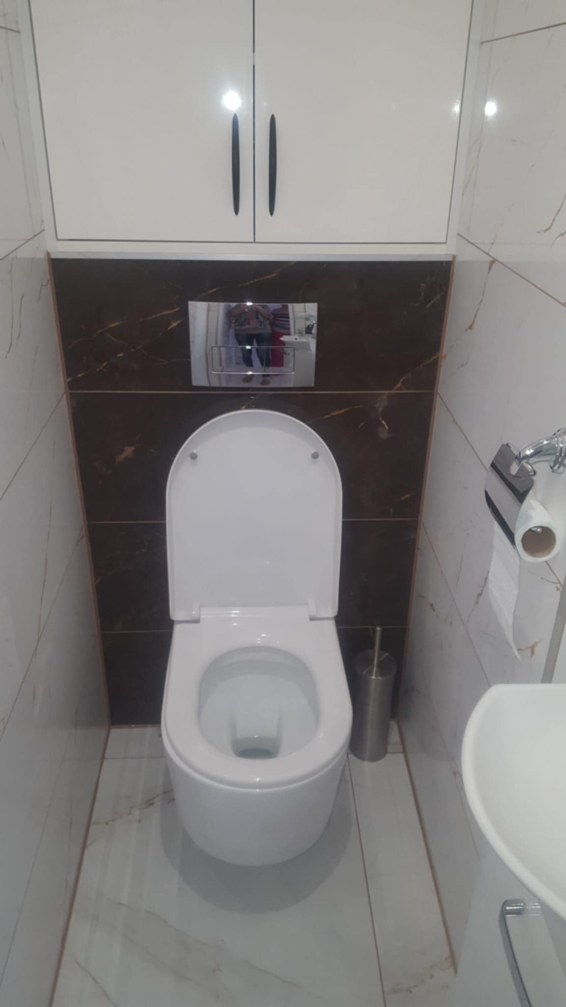 Nowoczesna toaleta wisząca z deską sedesową, zamontowana na tle brązowych płytek imitujących marmur, z szafką nad nią; widoczny również uchwyt na papier toaletowy i fragment umywalki.