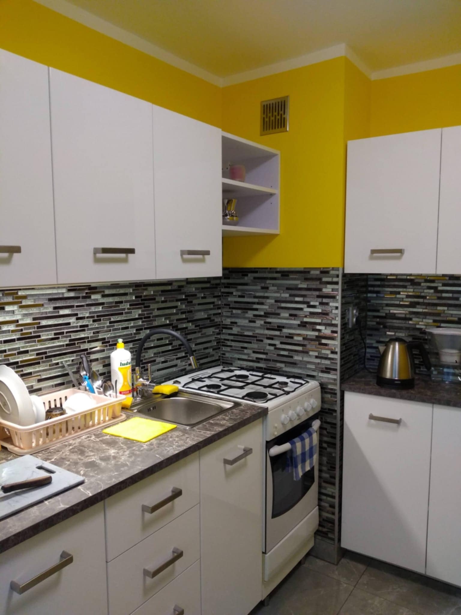 Nowoczesna kuchnia z białymi szafkami, mozaikową ścianą nad blatem i intensywnie żółtą ścianą w tle, widok na zlewozmywak, kuchenkę i akcesoria kuchenne.