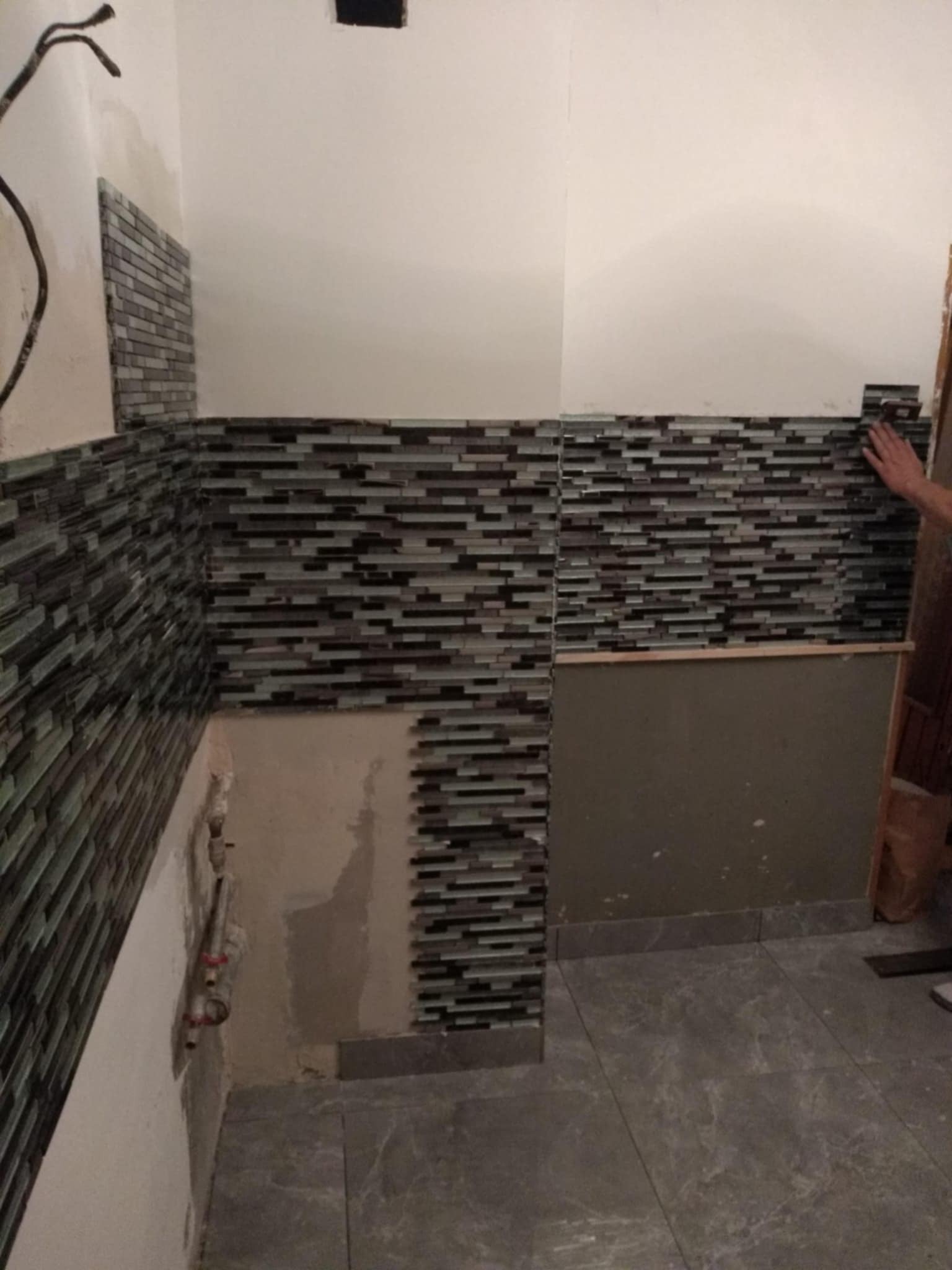 Montaż mozaiki z drobnych, poziomych płytek w odcieniach szarości i zieleni na ścianie łazienki, widoczne fragmenty instalacji hydraulicznej i niedokończone fragmenty ściany.