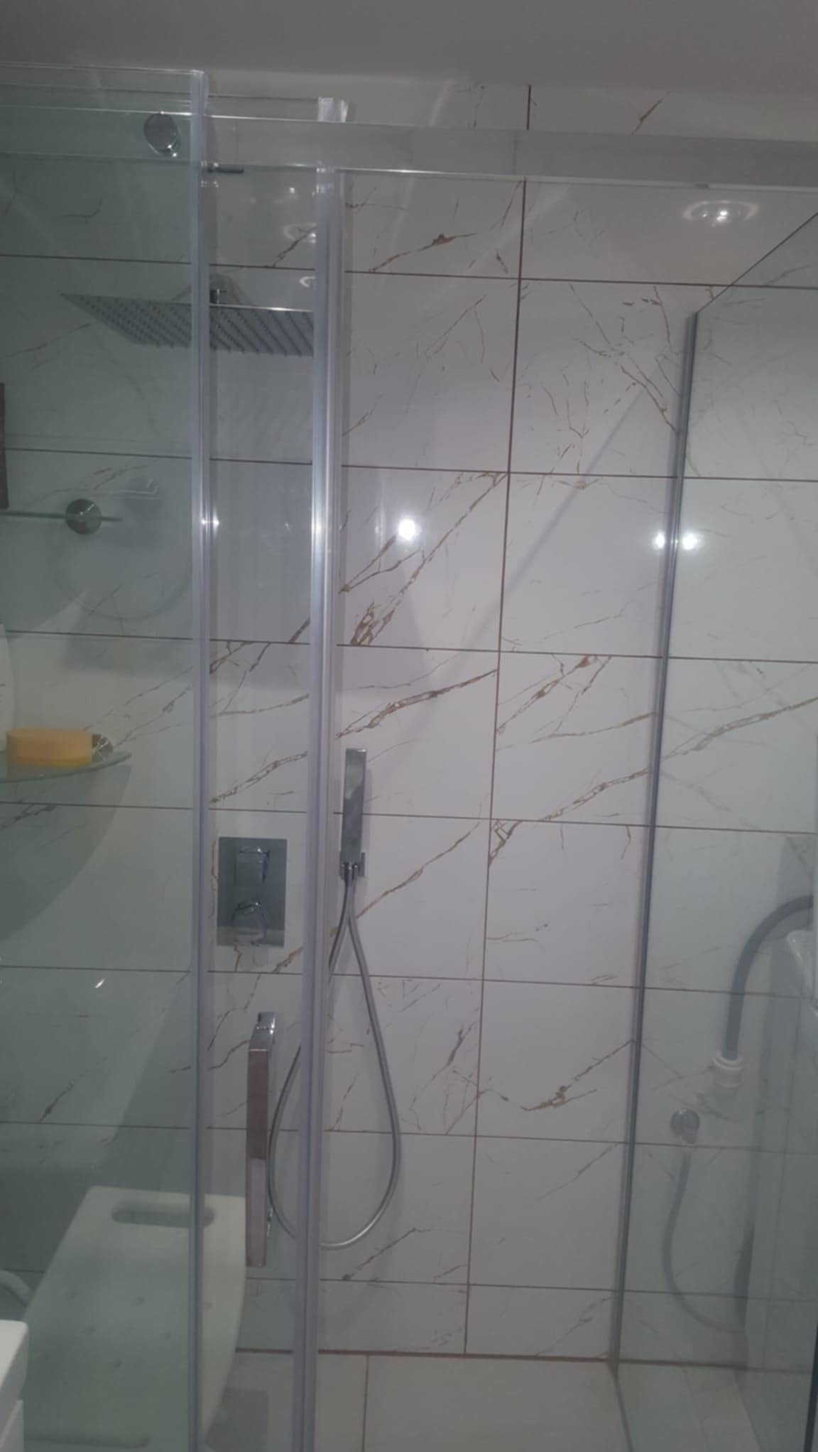 Nowoczesna kabina prysznicowa z deszczownicą i szklanymi drzwiami na tle białych płytek imitujących marmur z delikatnymi złotymi żyłkami, widoczny fragment toalety