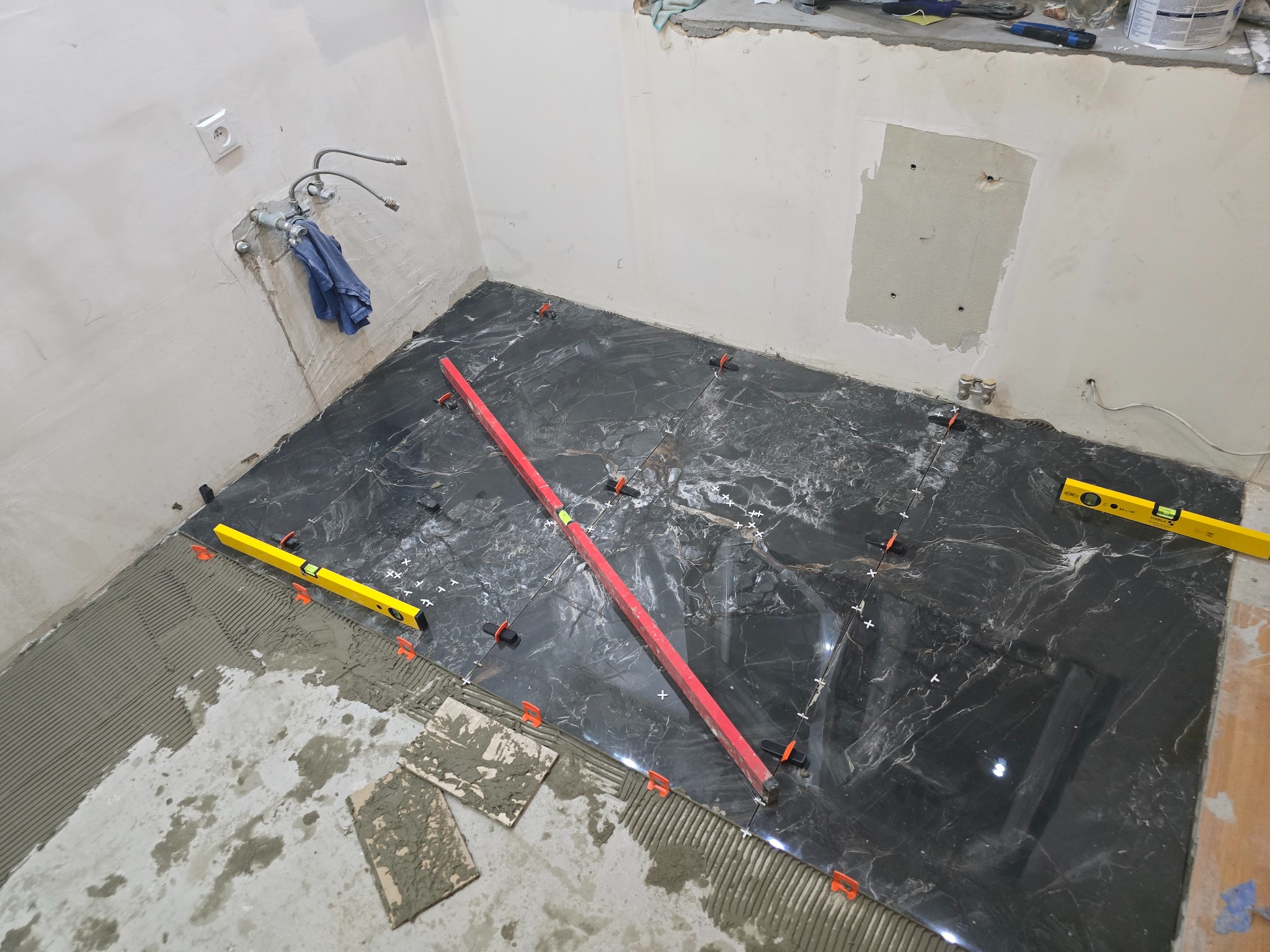 Układanie czarnych, marmurkowych płytek podłogowych w łazience, z użyciem poziomnic i systemu poziomowania. Widoczne przygotowane podłoże i fragmenty nieułożonych płytek.