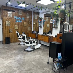 SanyRum - Wnętrze nowoczesnego salonu fryzjerskiego z drewnianymi panelami, neonem 'Fajnie, że jesteś', dwoma fotelami barberskimi i roślinami doniczkowymi.