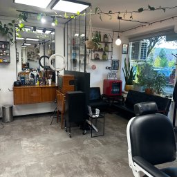 SanyRum - Wnętrze barbershopu w stylu retro: drewniane meble, rośliny pnące, czarne fotele, oświetlenie LED i dekoracyjny neon z napisem 'Darmowe napoje'.