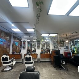 SanyRum - Wnętrze nowoczesnego salonu fryzjerskiego z drewnianą ścianą, neonem i roślinami. Widoczne stanowiska z fotelami fryzjerskimi i lustrami.