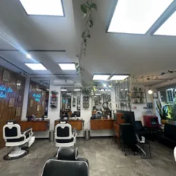 Wnętrze nowoczesnego salonu fryzjerskiego z drewnianą ścianą, neonem i roślinami. Widoczne stanowiska z fotelami fryzjerskimi i lustrami.