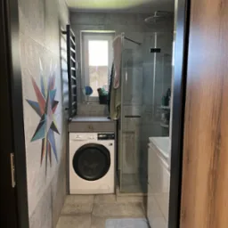 Szara łazienka z kabiną prysznicową i pralką Electrolux. Na ścianie dekoracja w kształcie gwiazdy. Nowoczesne wnętrze z szarymi płytkami.