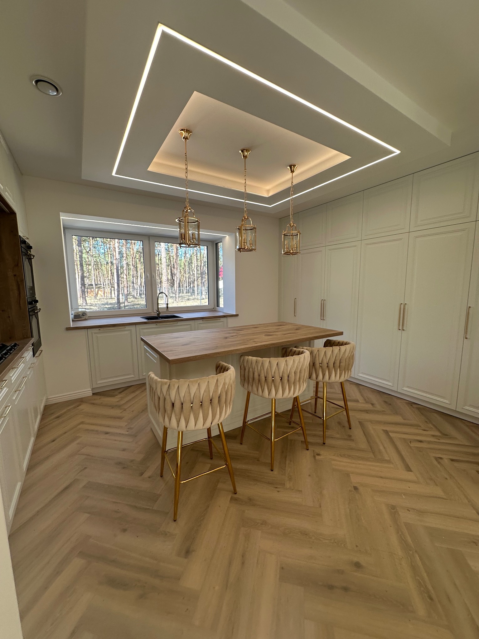 Jasna kuchnia z drewnianym blatem wyspy i trzema krzesłami na złotych nogach. Nad wyspą trzy złote lampy. Parkiet w jodełkę. Okno z widokiem na las.