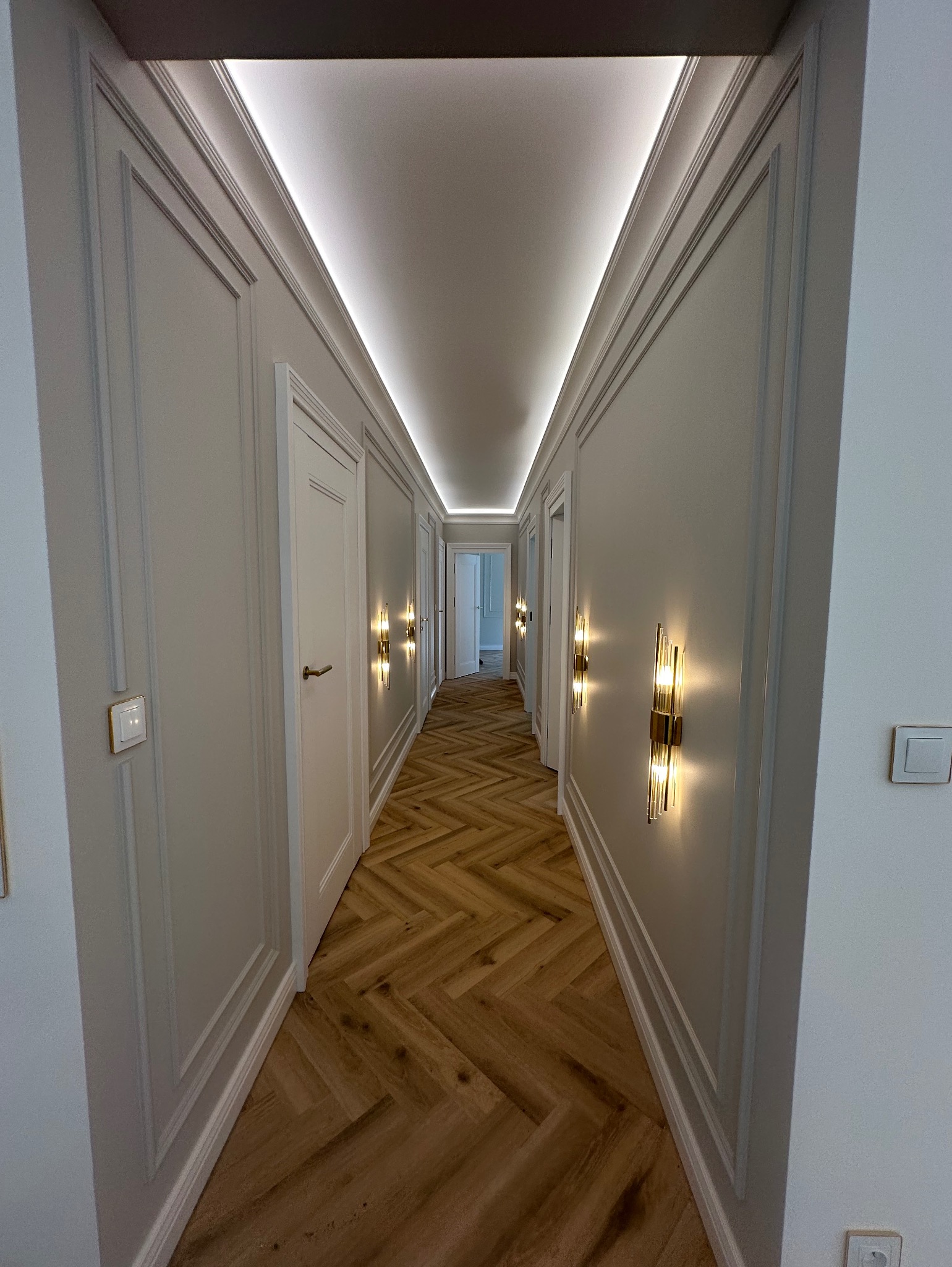 Korytarz w stylu klasycznym z parkietem w jodełkę, oświetleniem LED na suficie i ozdobnymi kinkietami. Białe ściany z dekoracyjnymi listwami.